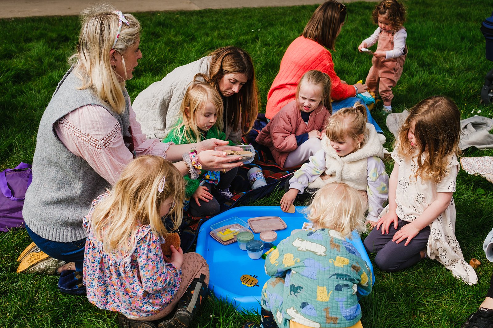 Group of children and adults sitting on grass during a play activity with toys and art supplies, in an outdoor setting.