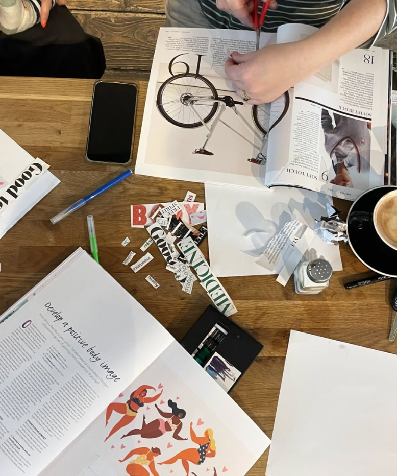 A wooden table covered with magazines, a smartphone, pens, and scattered paper cutouts. One magazine shows an illustration of women dancing, and another open magazine displays a photo of a bicycle. There is a cup of coffee, a small salt shaker, and a black pen.