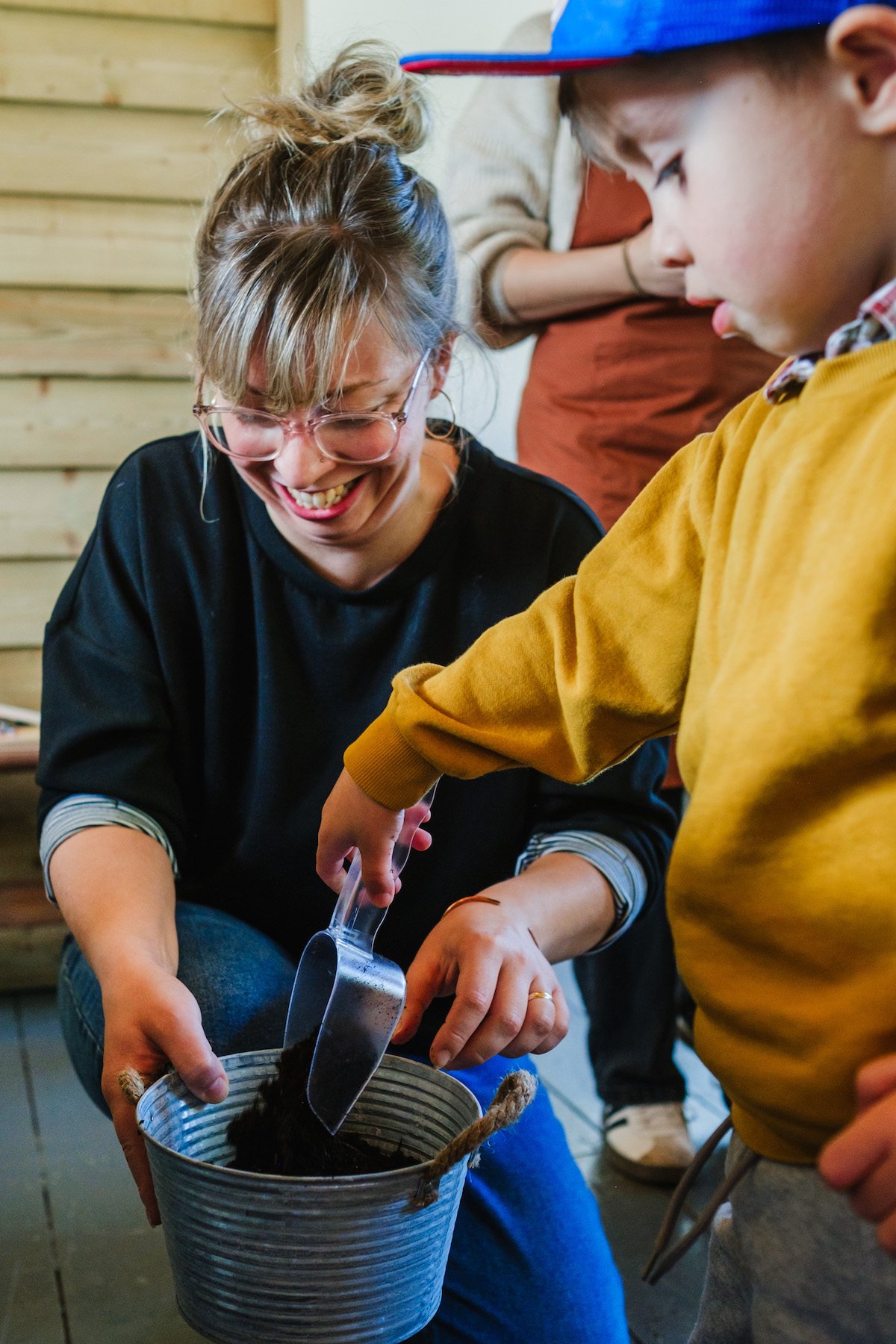 A woman smiling and wearing glasses, helping a young boy plant a seed into a small container, using a trowel. Another person stands in the background with arms crossed, observing. The scene takes place in a room with wooden walls.