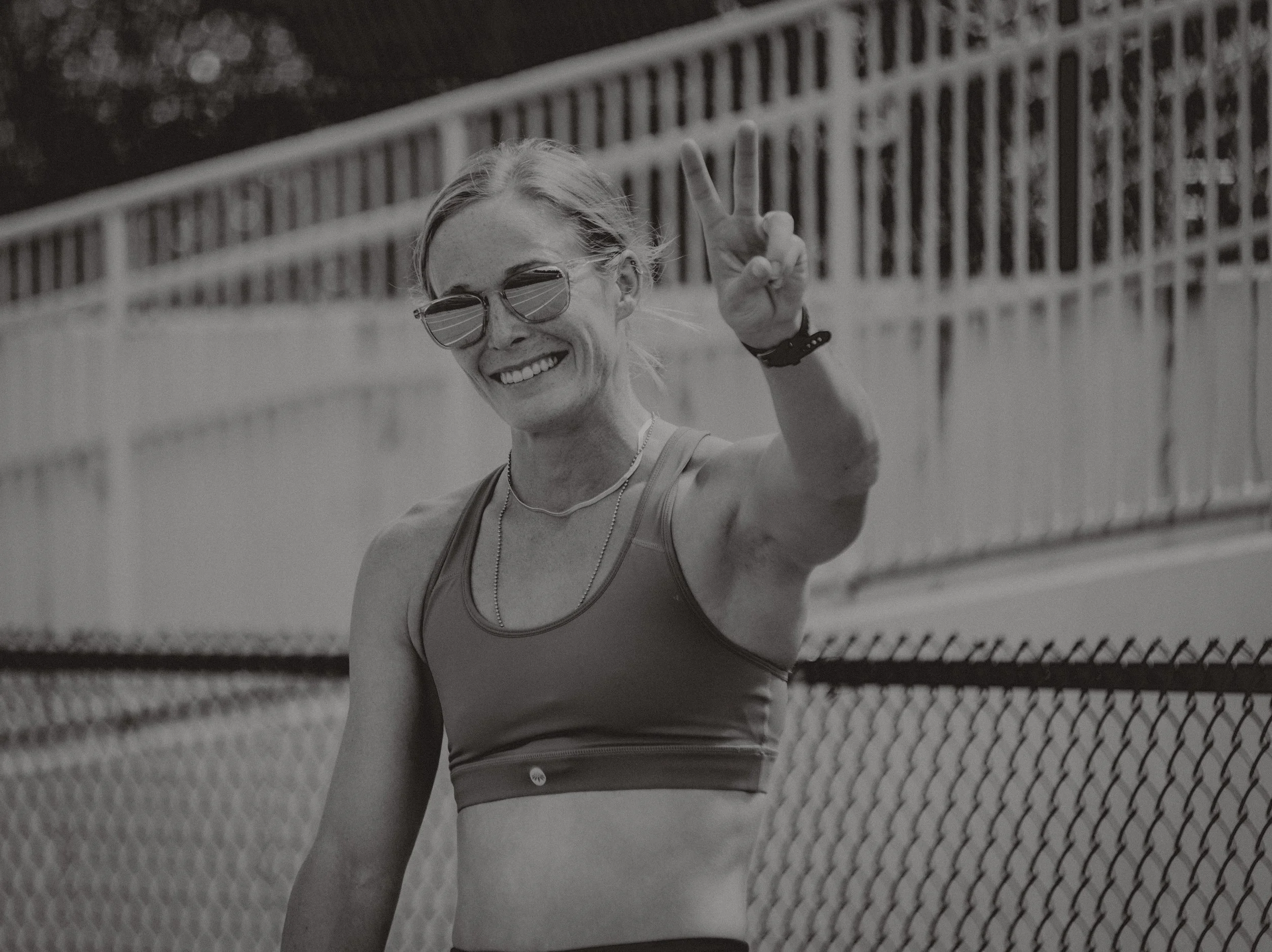 A smiling woman wearing sunglasses, a sports bra, and a necklace, making a peace sign with her right hand on a bridge.