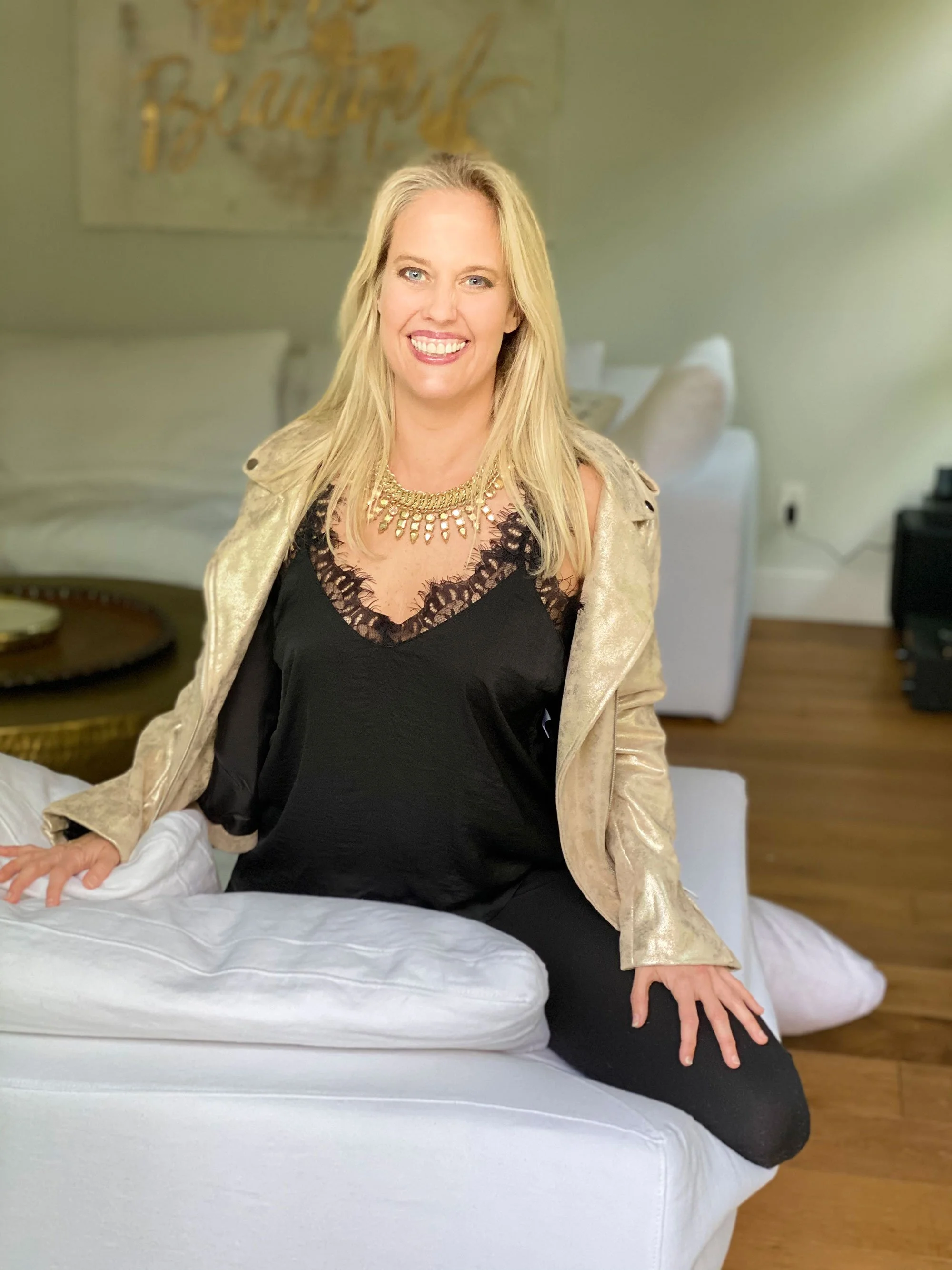 A smiling blonde woman in a black top with lace detail and a gold jacket, sitting on a white couch in a living room.
