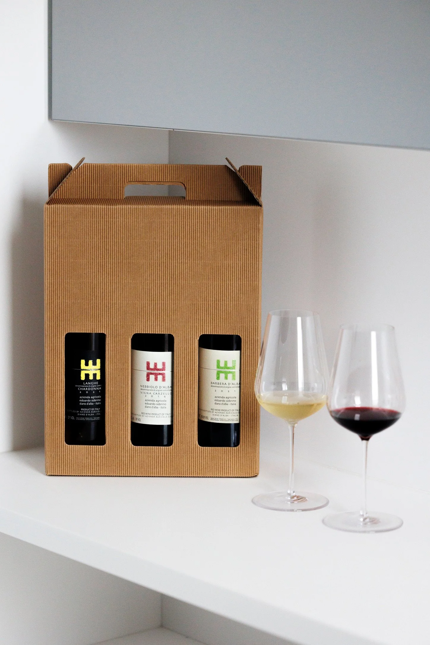 Een kartonnen wijnkistje met drie flessen wijn, naast twee wijnglazen met witte en rode wijn op een witte tafel in een minimalistische interieur.