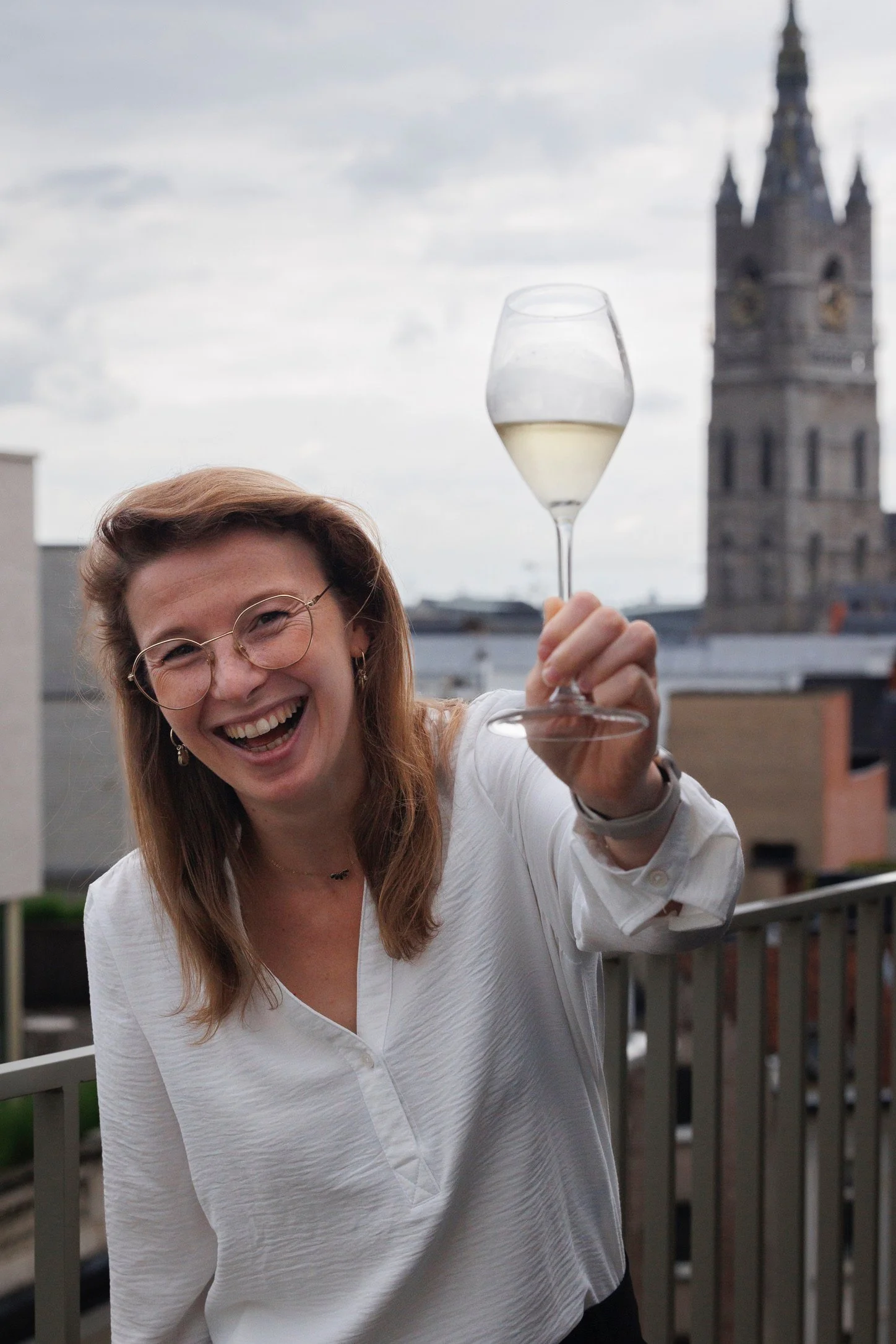 Een lachende vrouw met bruin halflang haar, glazen bril en sieraden, die een glas witte wijn omhoog houdt op een balkon met uitzicht op een kerk of toren in de achtergrond.