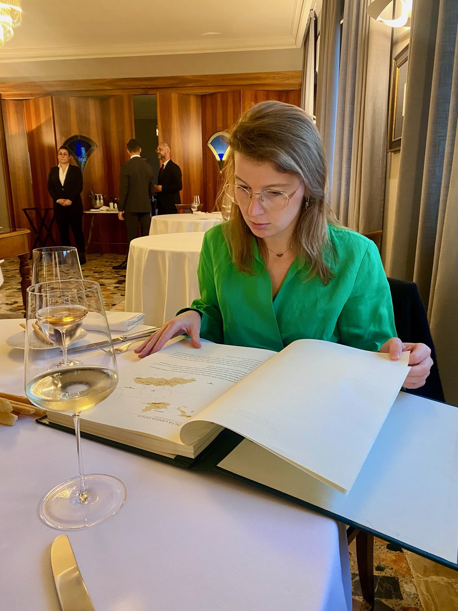 Vrouw leest een groot boek in een formeel restaurant met twee glazen witte wijn voor haar, omringd door witte tafelkleden, in een elegante ruimte met houten wanden en gordijnen.