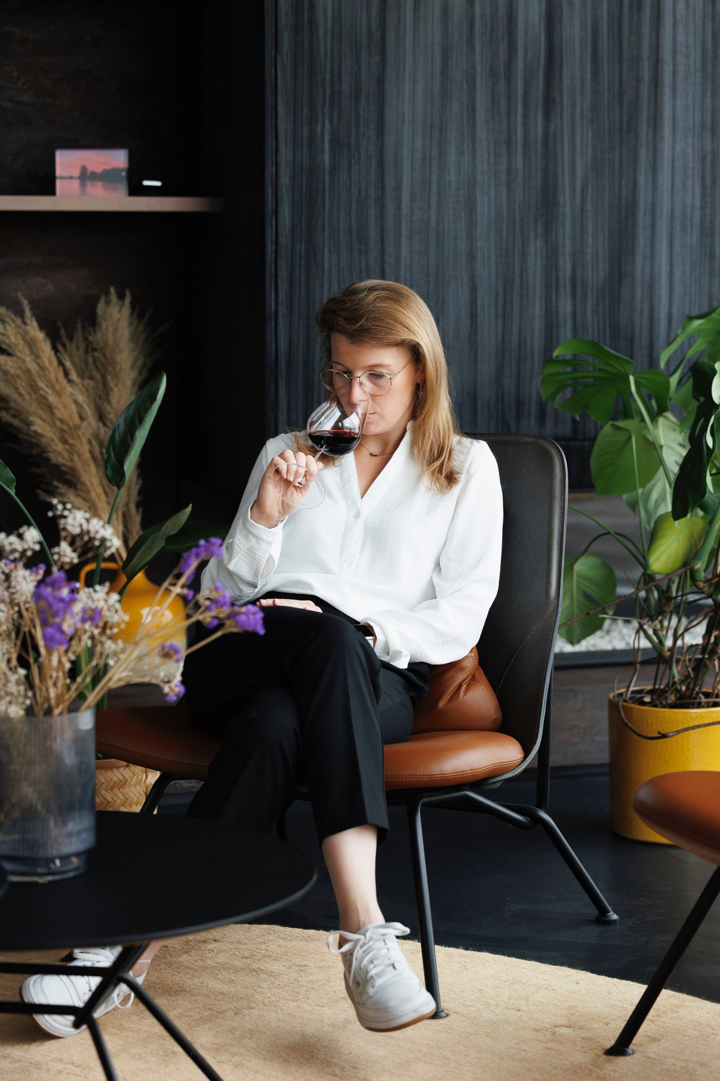 Vrouw die rode wijn drinkt terwijl ze op een stoeltje zit in een modern interieur met planten.