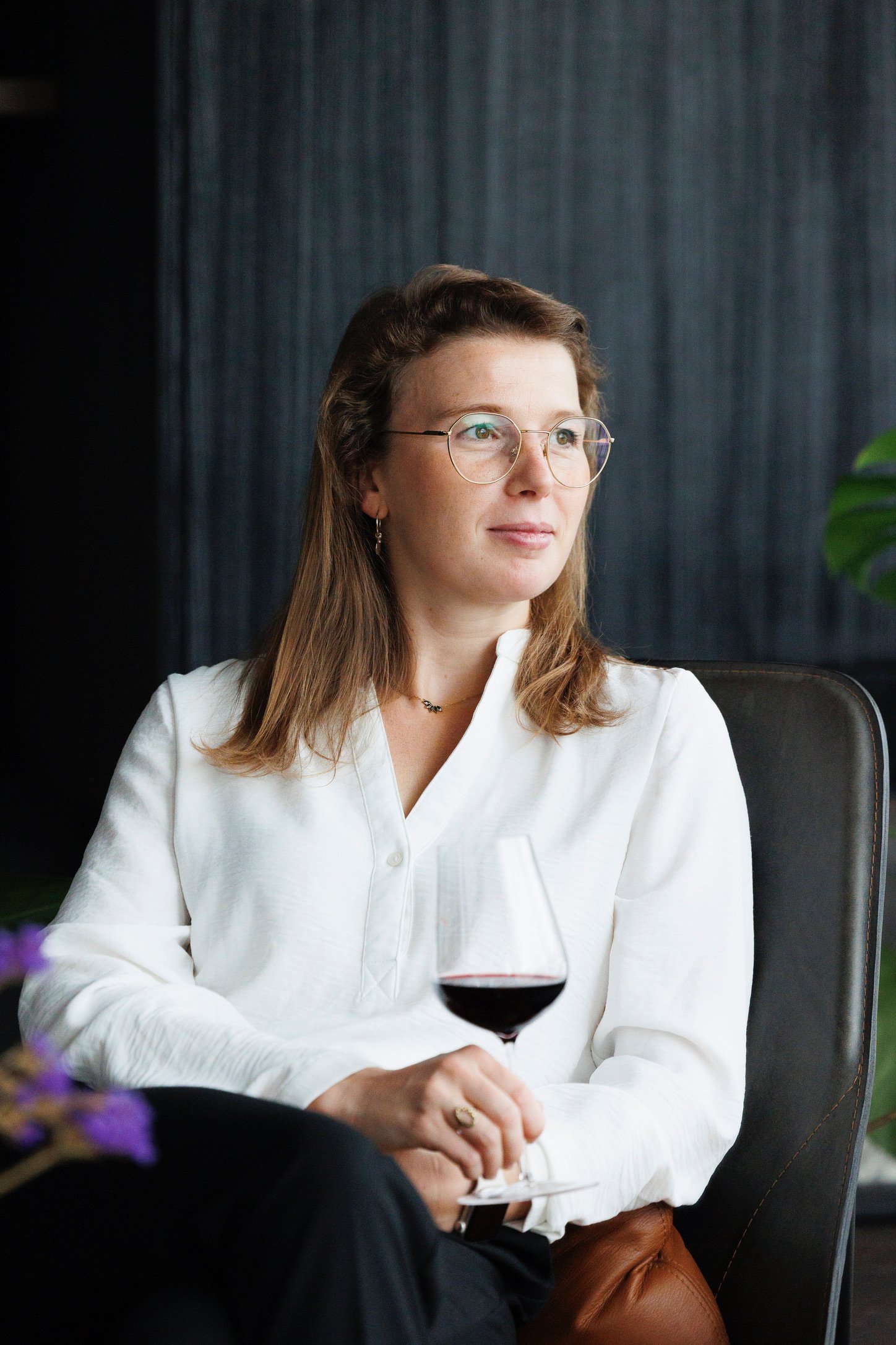 Vrouw met bruin haar, bril en witte blouse zit met een glas rode wijn in haar hand, kijkt uit het raam, op de achtergrond een donkere muur en een groene plant.