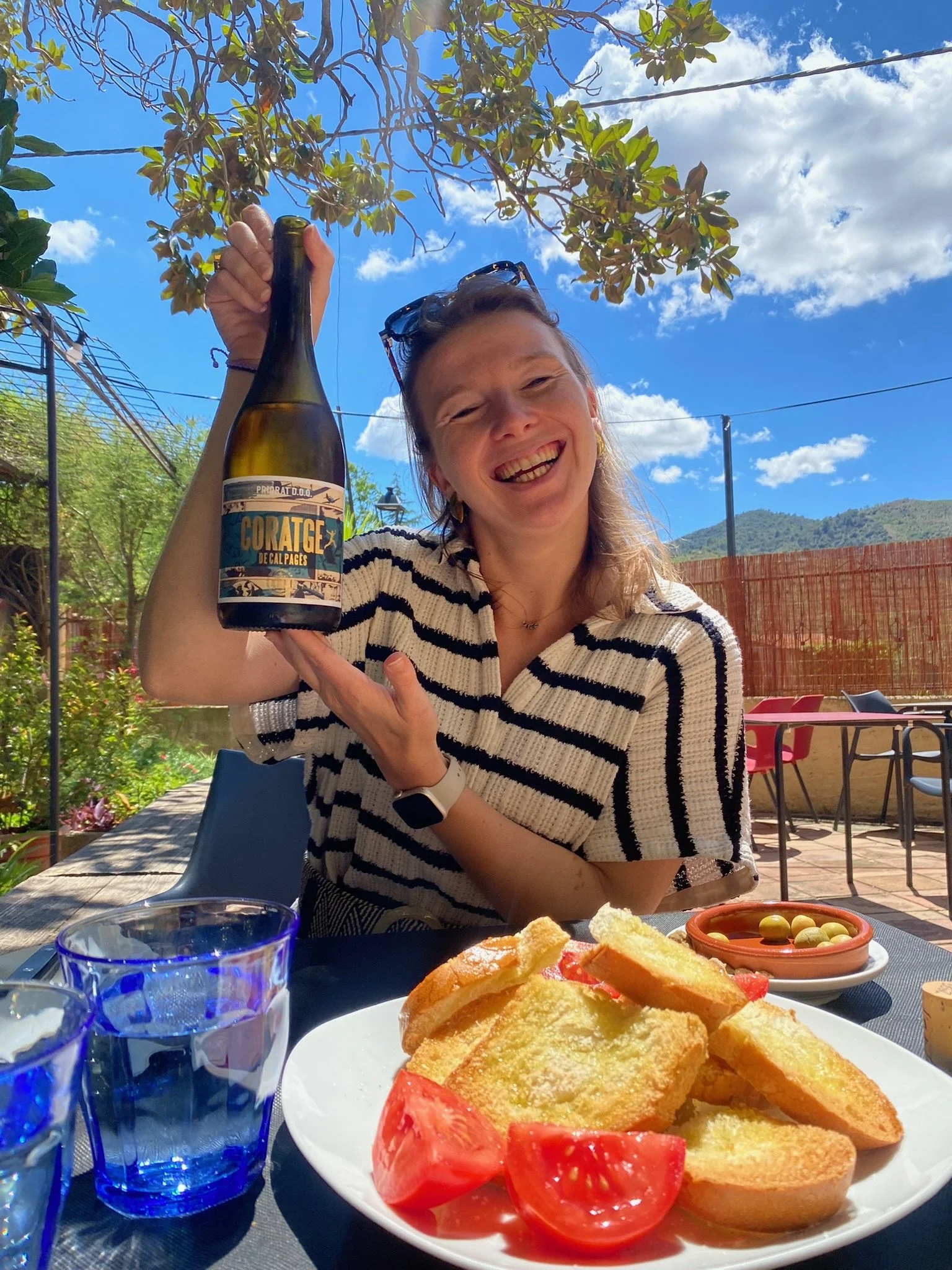 Vrolijke vrouw met zonnebril op haar hoofd die een fles wijn vasthoudt, zittend aan een tafel met tapas en brood, buiten onder een boom bij een zonnige dag.