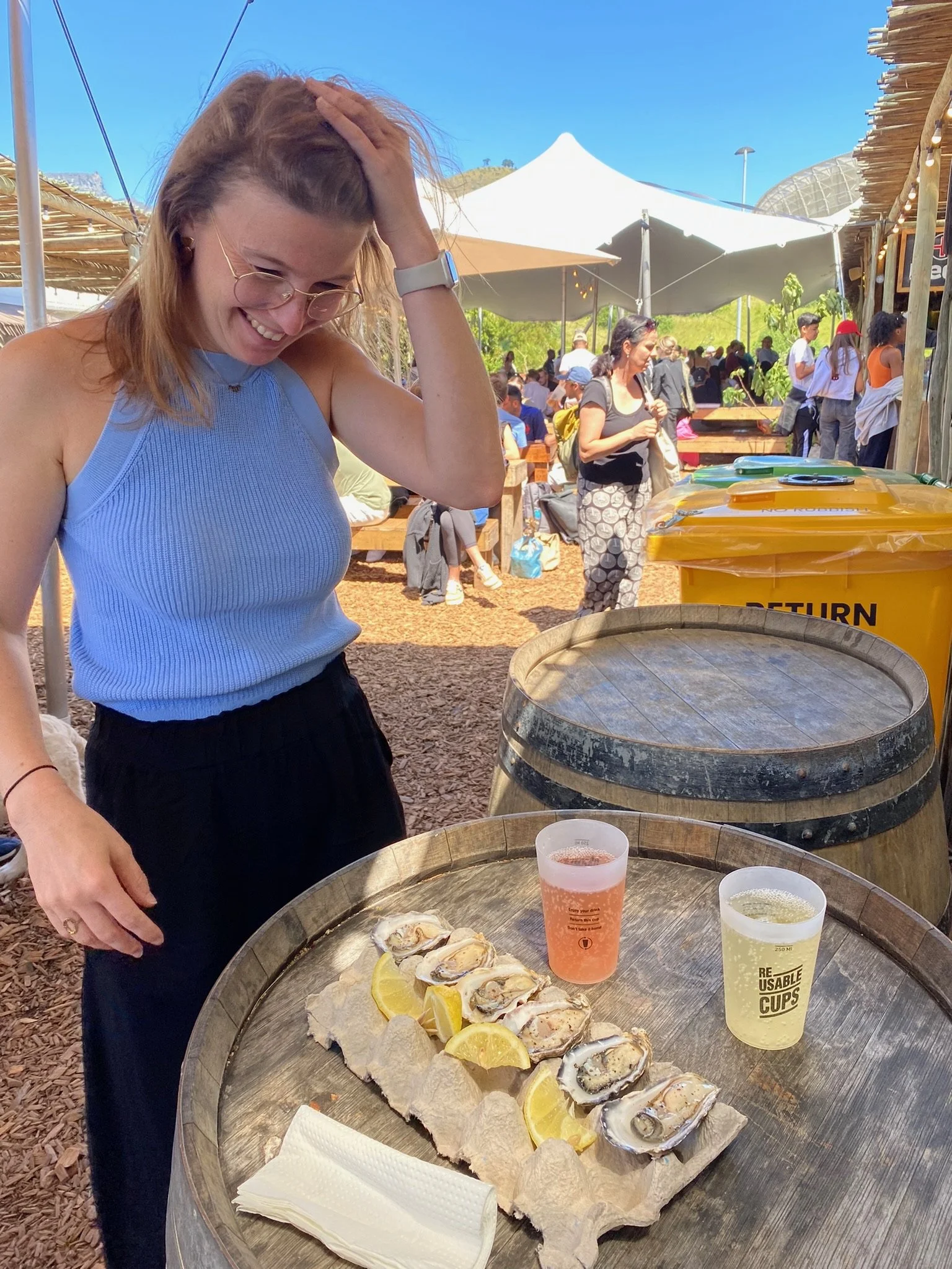 Een vrouw staat lachen naast een baril met een schaal met oesters, citroenpartjes en twee drankjes op de tafel. Op de achtergrond is een drukke outdoor markt met veel mensen, parasols en houten winkeltjes.