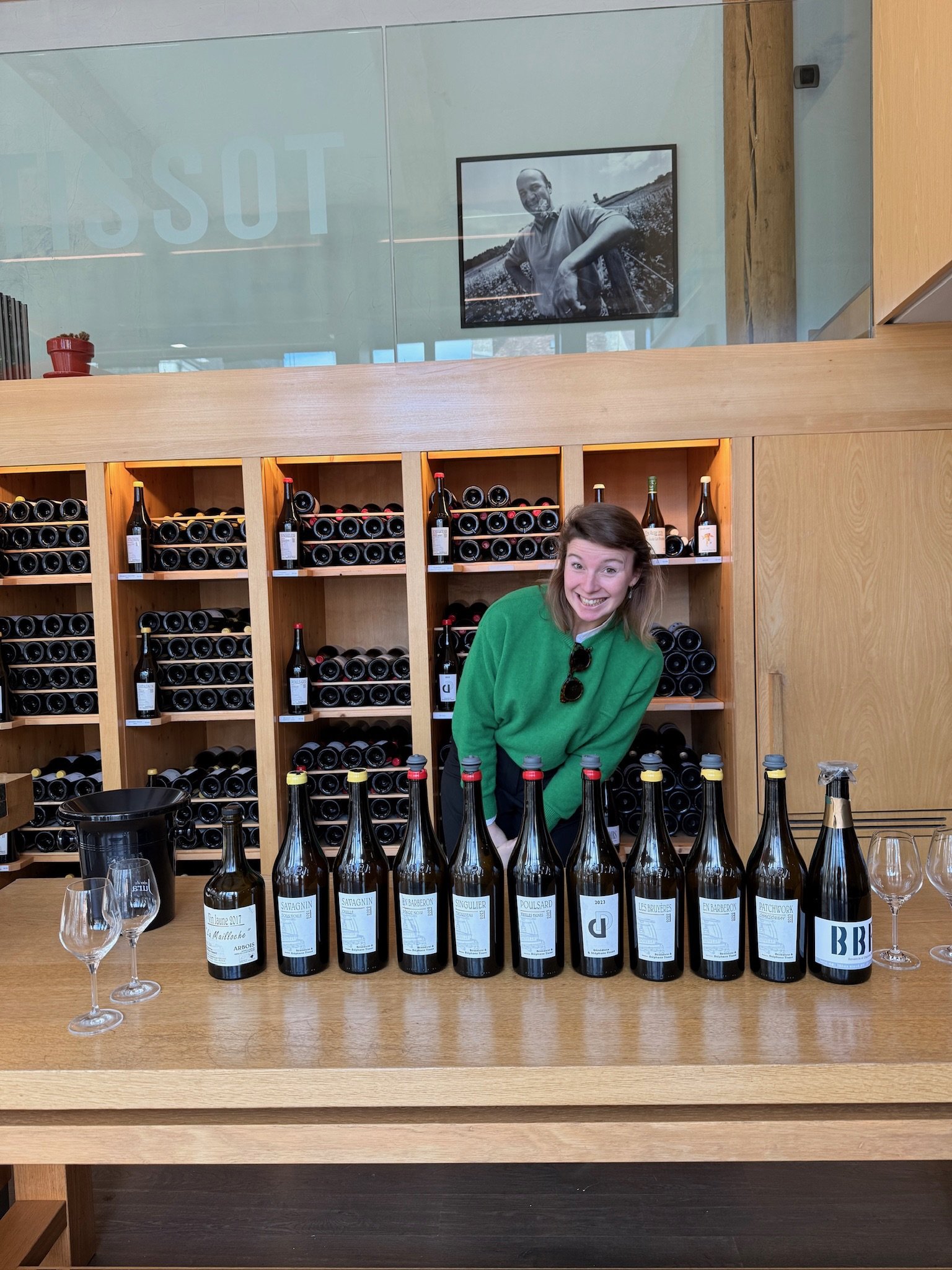 Vrouw met bruin haar en groene trui die glimlacht achter een tafel met 12 wijnflessen en twee lege wijnglazen in een wijnwinkel of proeflokaal, met wijnrekken op de achtergrond en een zwarte en witte foto op de muur.