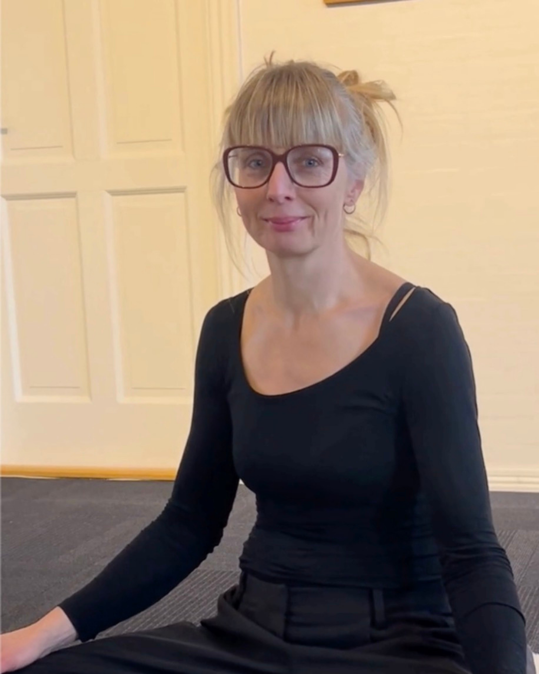 Kvindelig person med blond hår, sorte briller, siddende på pude, i et indendørs miljø med beige baggrund og gul lampeslynge.