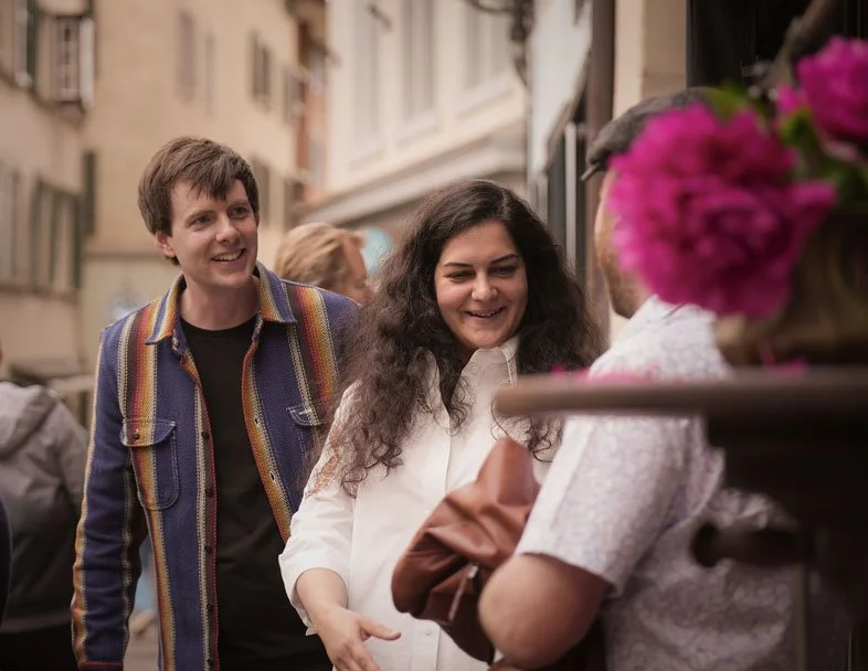 Drei Menschen unterhalten sich draußen in einer Stadtstraße. Zwei Frauen und ein Mann lachen und sprechen miteinander, im Hintergrund weitere Passanten.