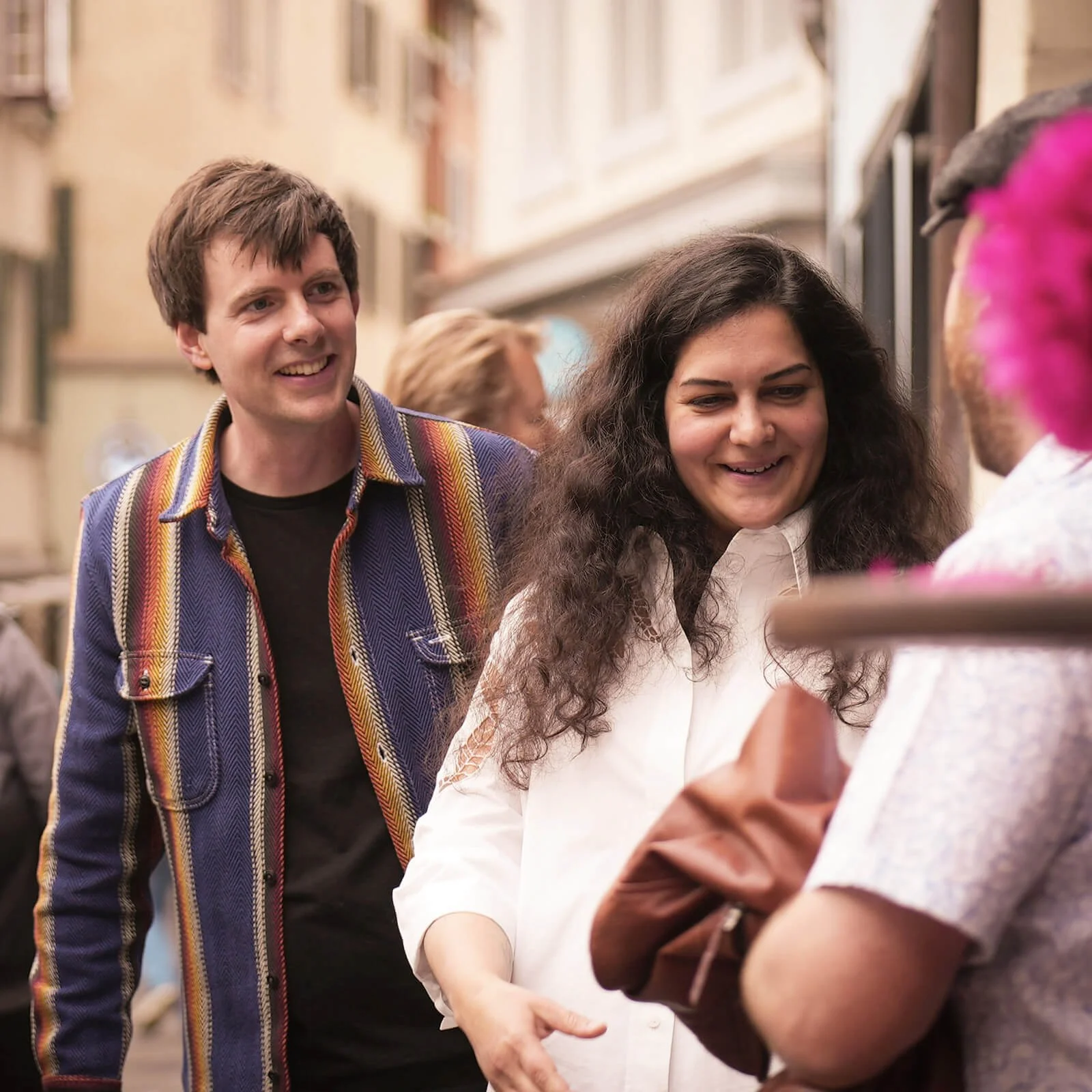 Drei junge Menschen lächeln und sprechen in einer belebten Straße mit alten Gebäuden.