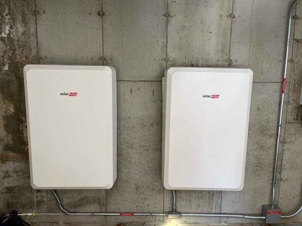 The image shows a home battery storage system installed in a basement, positioned next to electrical panels, highlighting a modern approach to energy management. This setup supports solar energy production by storing excess energy.