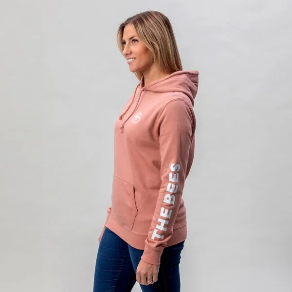 on model photography of a woman with long blonde hair wearing a pink sportwear hoodie with white text on the sleeve that reads "THE BEES" and blue jeans, standing against a plain light gray background.