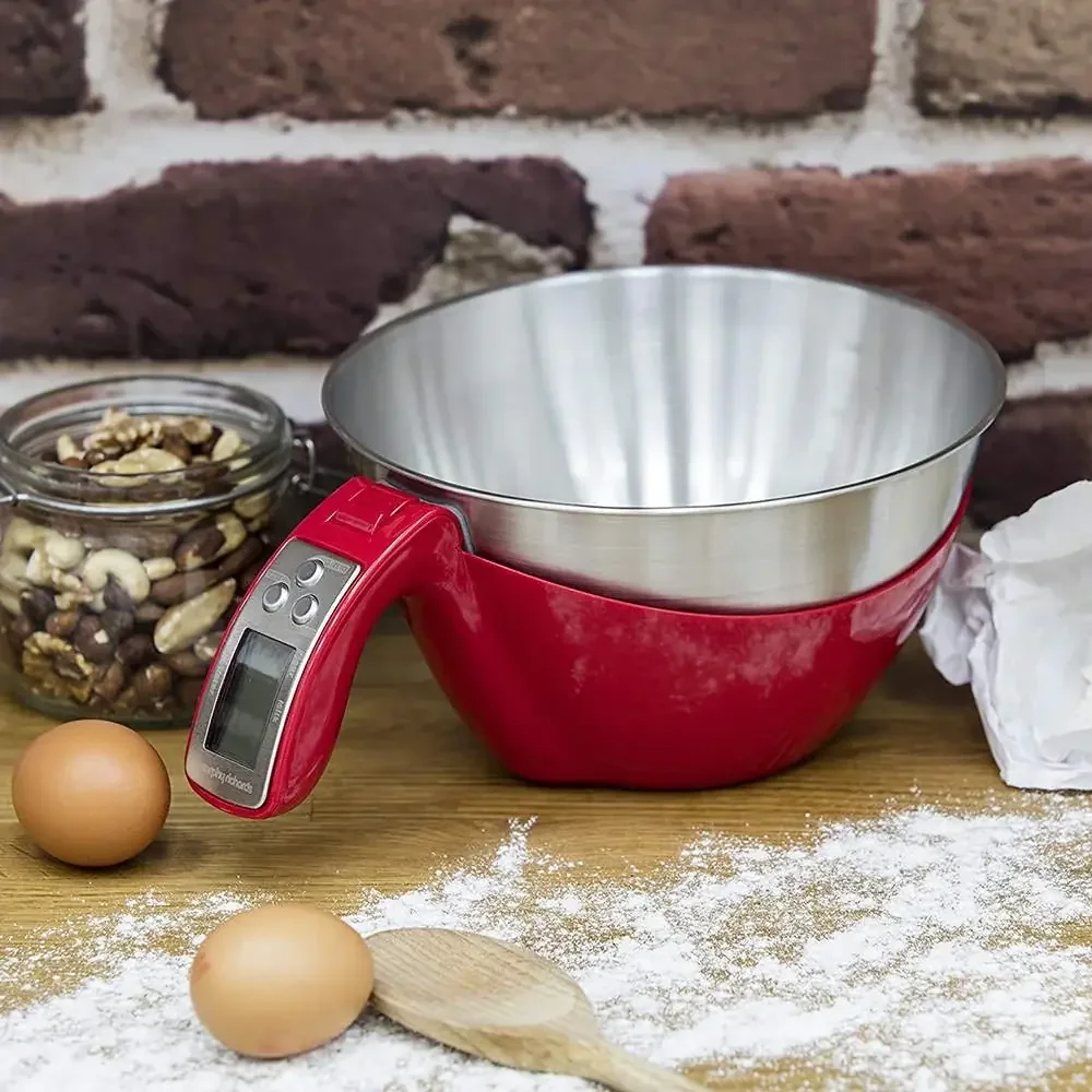 lifestyle image of red stand mixer with empty metal mixing bowl, two eggs, a jar of mixed nuts, a wooden spoon, and scattered flour on a wooden surface against a brick wall.