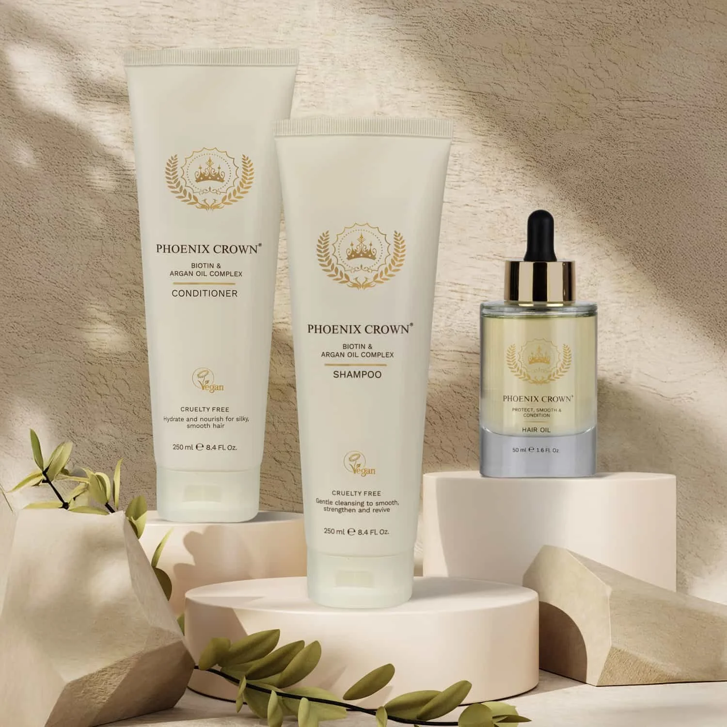 Display of three Phoenix Crown hair care products: conditioner, shampoo, and hair oil, arranged on white blocks with decorative green leaves and a textured beige background.