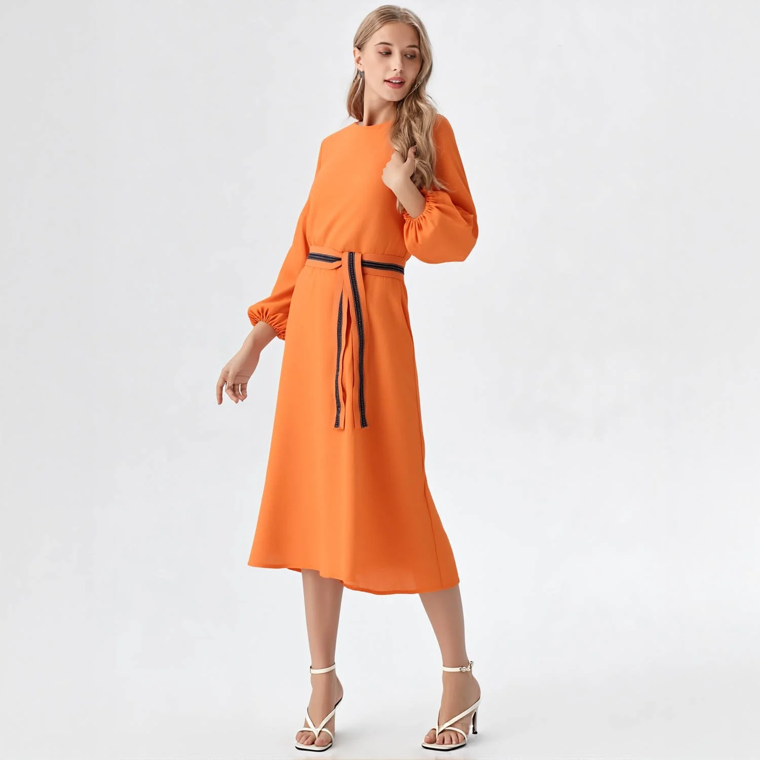 A female virtual on model in an orange dress with long sleeves, a black belt, and white heels, standing against a plain white background.