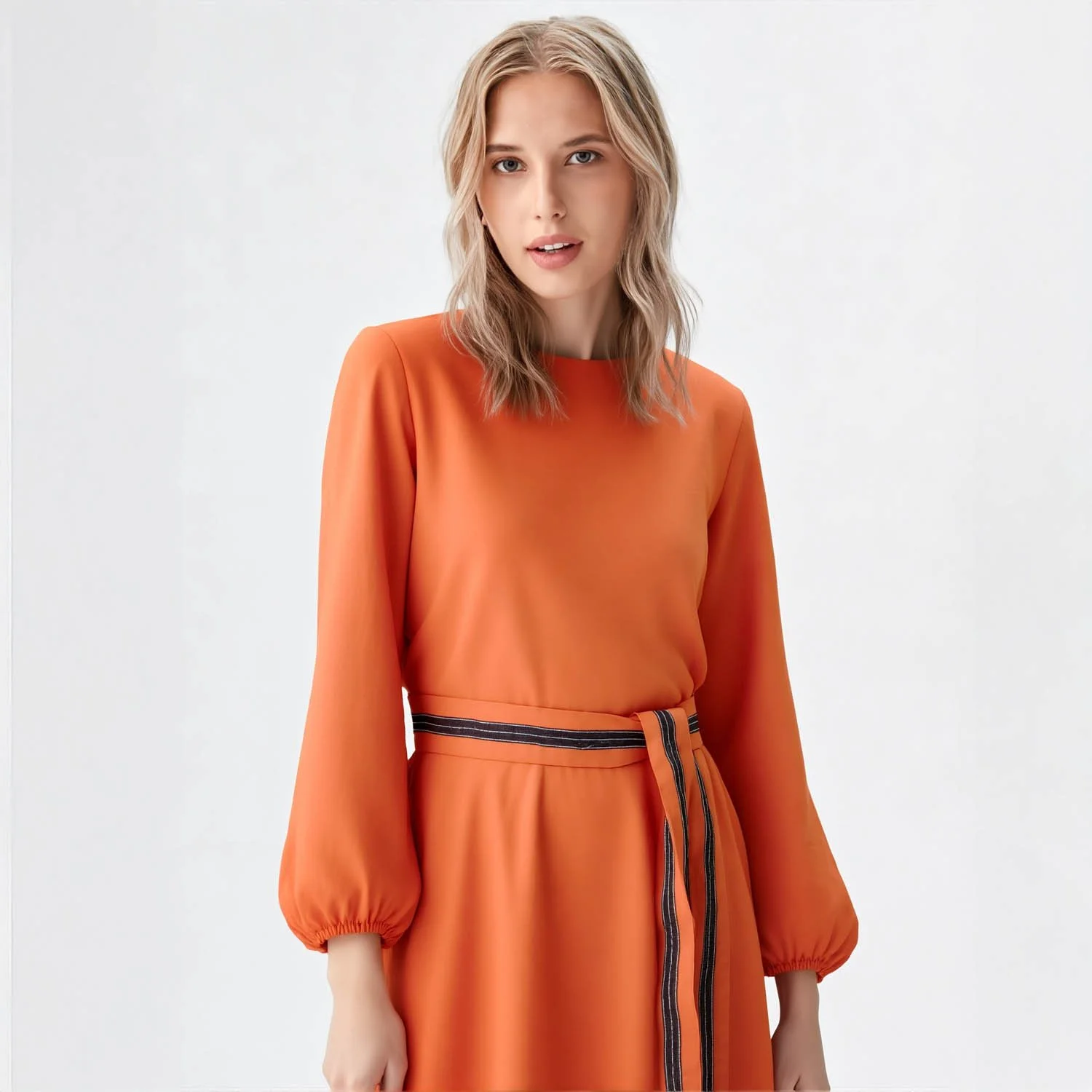 Virtual young female with blonde hair wearing an orange long-sleeve dress with a belt, standing against a plain white background.