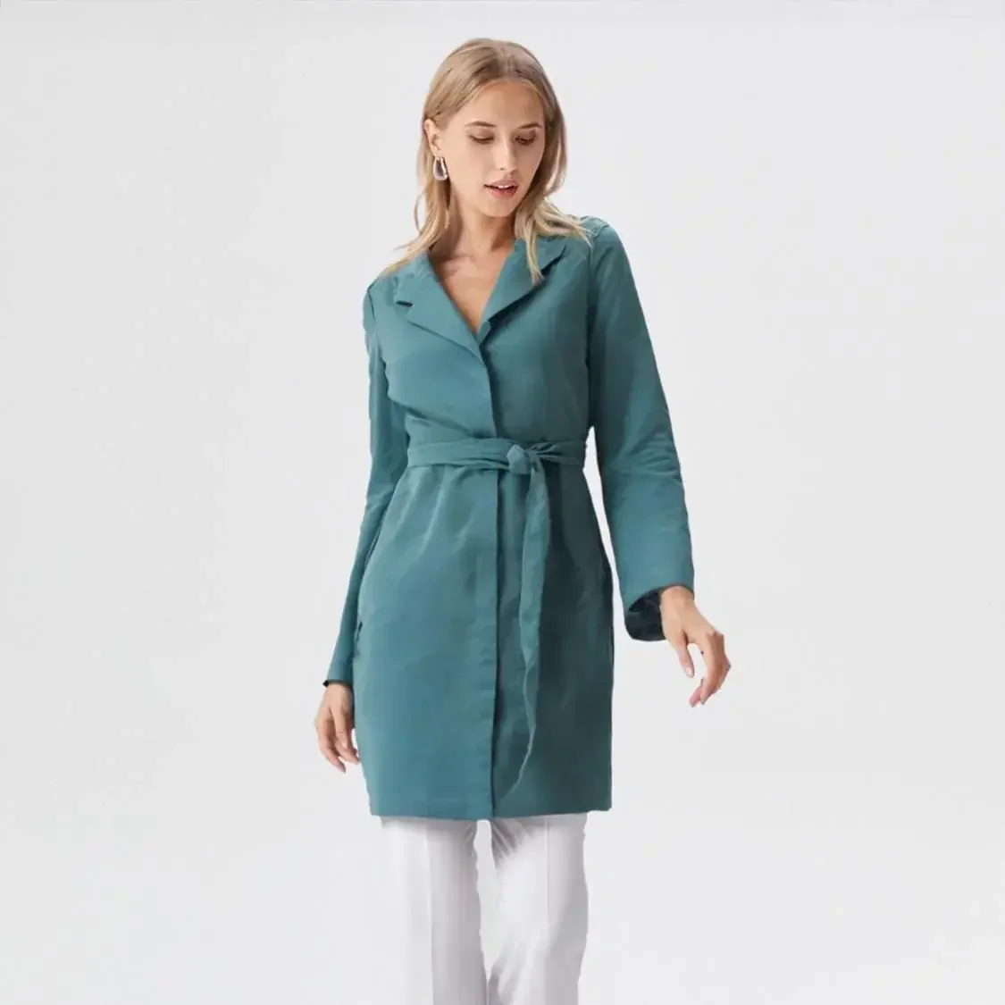 A virtual on model woman wearing a teal, long-sleeve, belted coat and white pants, standing against a plain background.