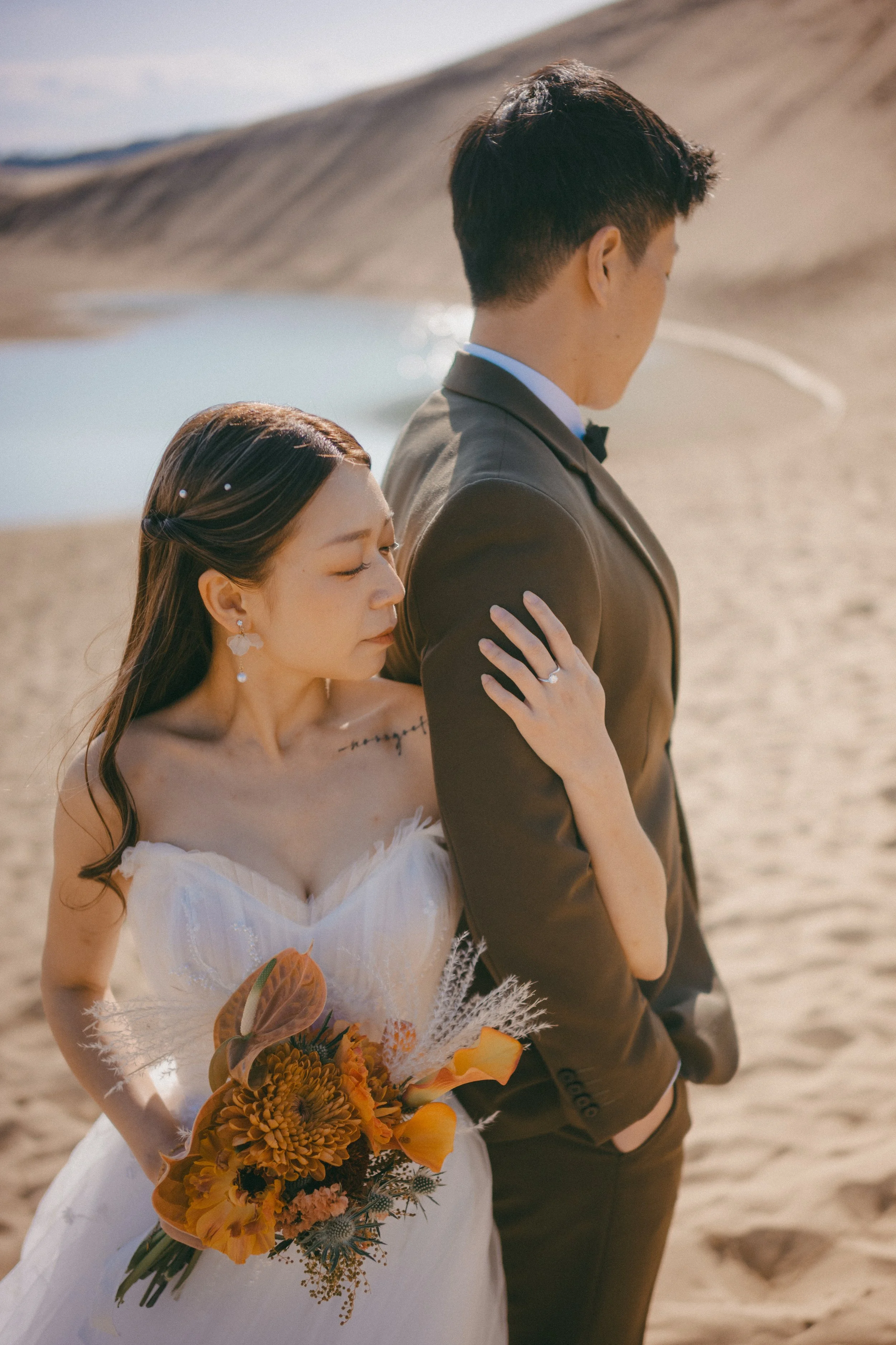 A bride and groom standing on a sandy beach, with the bride holding a bouquet of orange and yellow flowers. The bride has long hair and is wearing a white wedding dress, while the groom is in a dark suit. The woman is gently resting her hand on the m