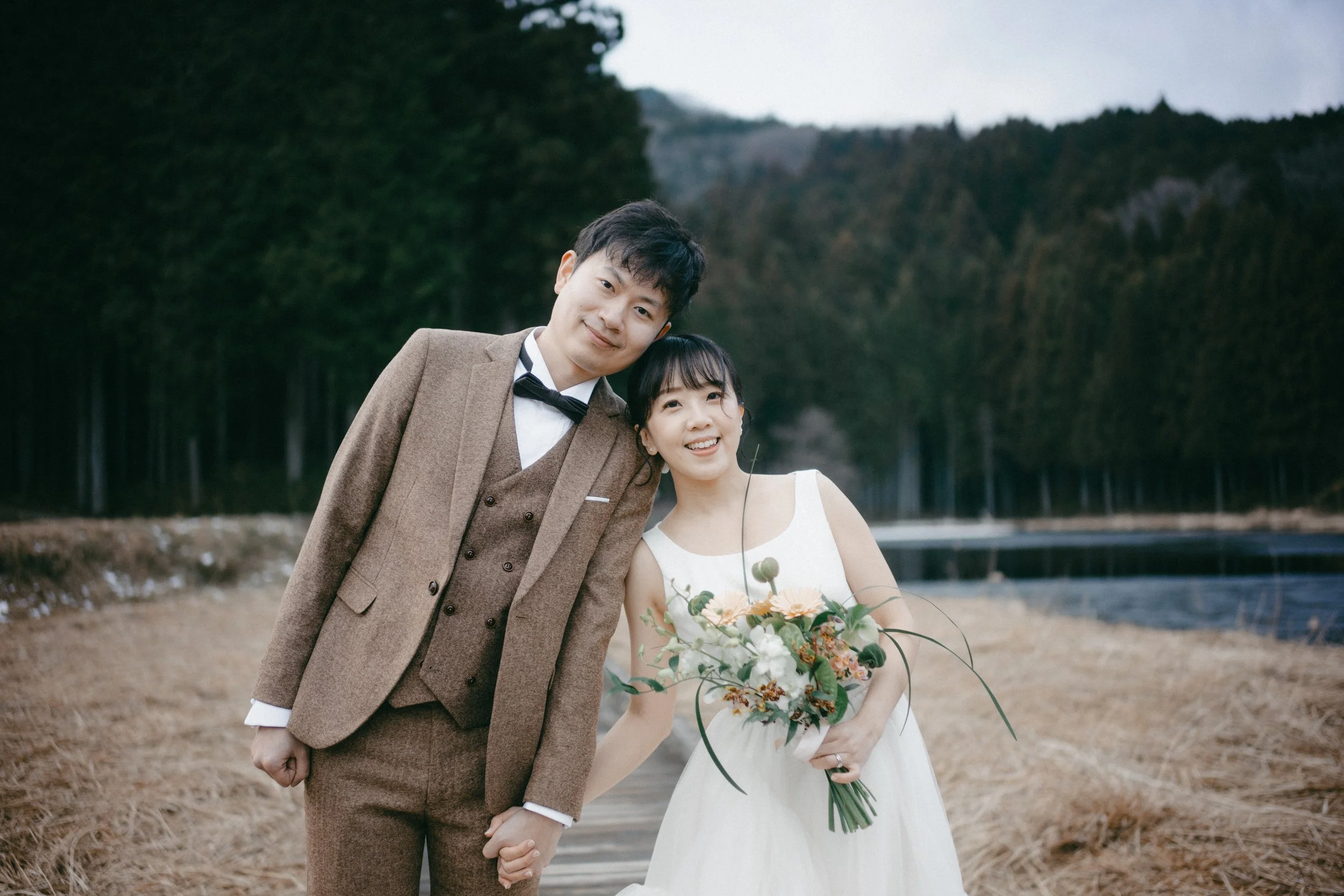 A newlywed couple standing outdoors near a body of water with a forest backdrop, holding hands and smiling. The groom is dressed in a brown suit with a bow tie, and the bride wears a white wedding gown holding a bouquet of flowers.