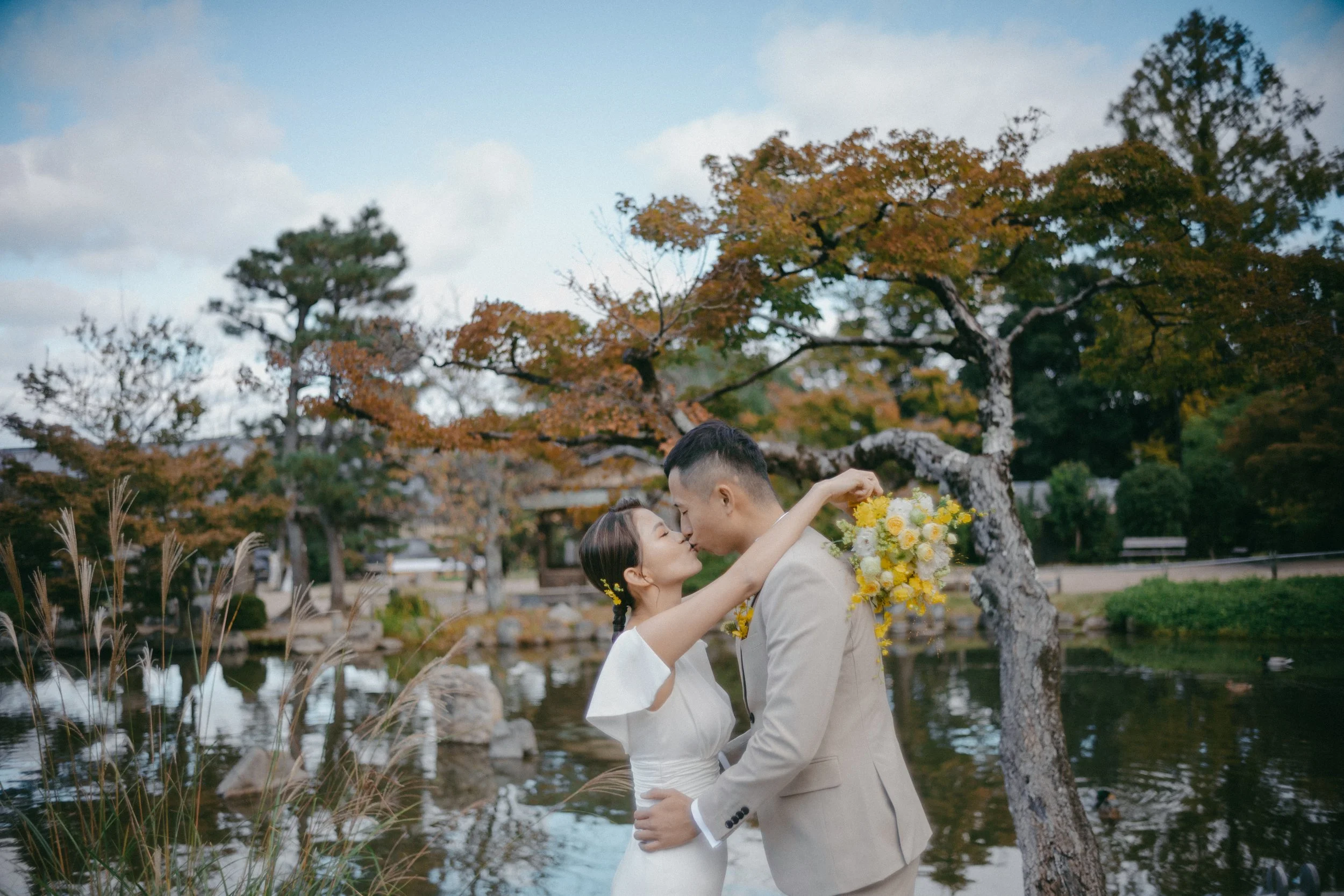 A couple kissing near a pond surrounded by autumn trees, one holding a floral bouquet.