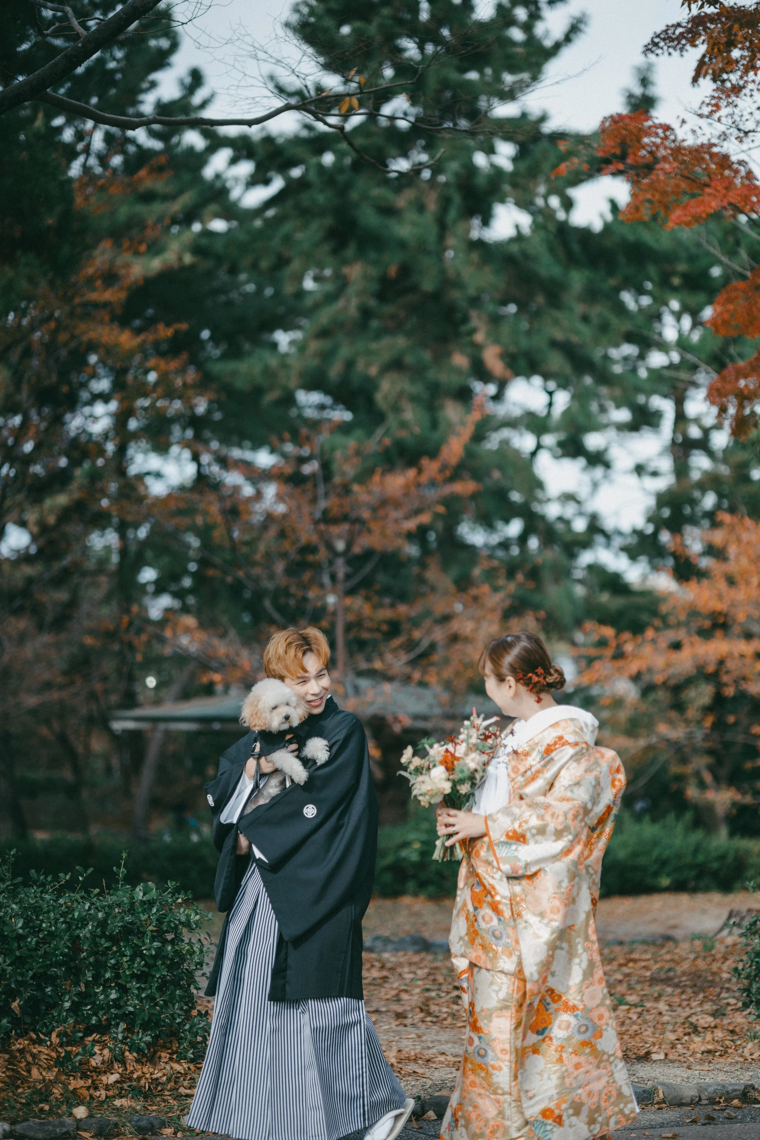A couple dressed in traditional Japanese kimonos outdoors with a small dog, surrounded by trees with autumn foliage.