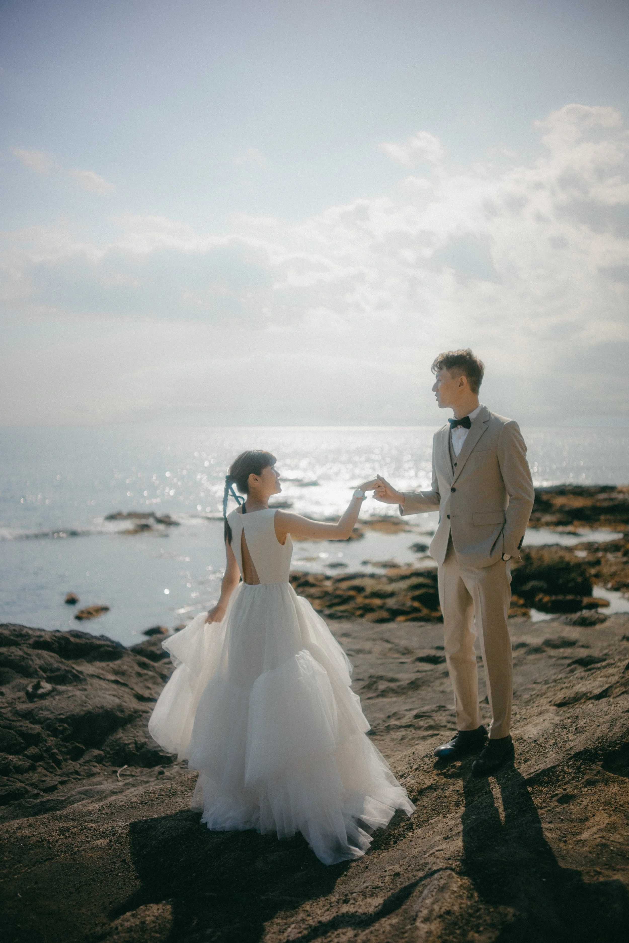 A couple dressed in wedding attire holding hands on a rocky beach with the ocean and sky in the background.