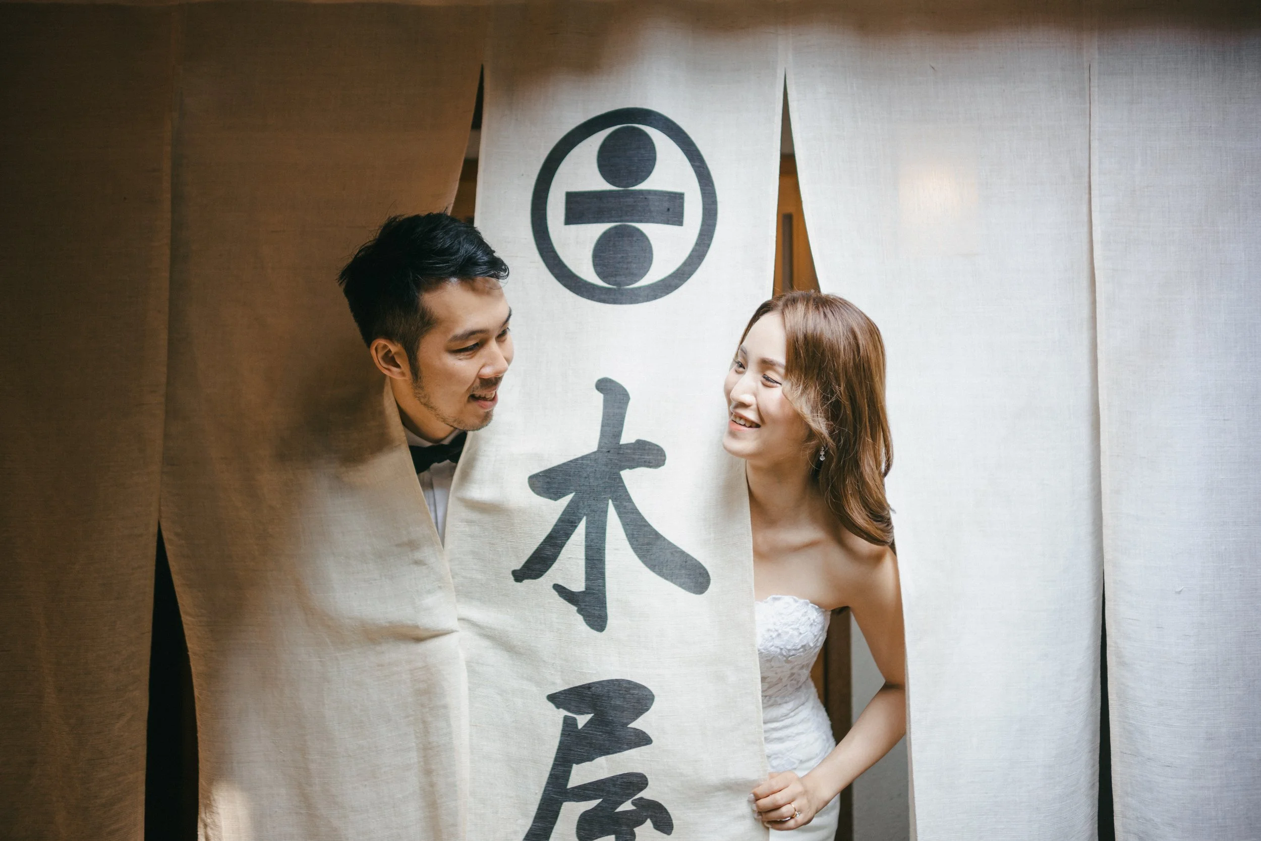 A couple in wedding attire peeking through traditional Japanese noren curtains with Japanese characters, smiling and looking at each other warmly.