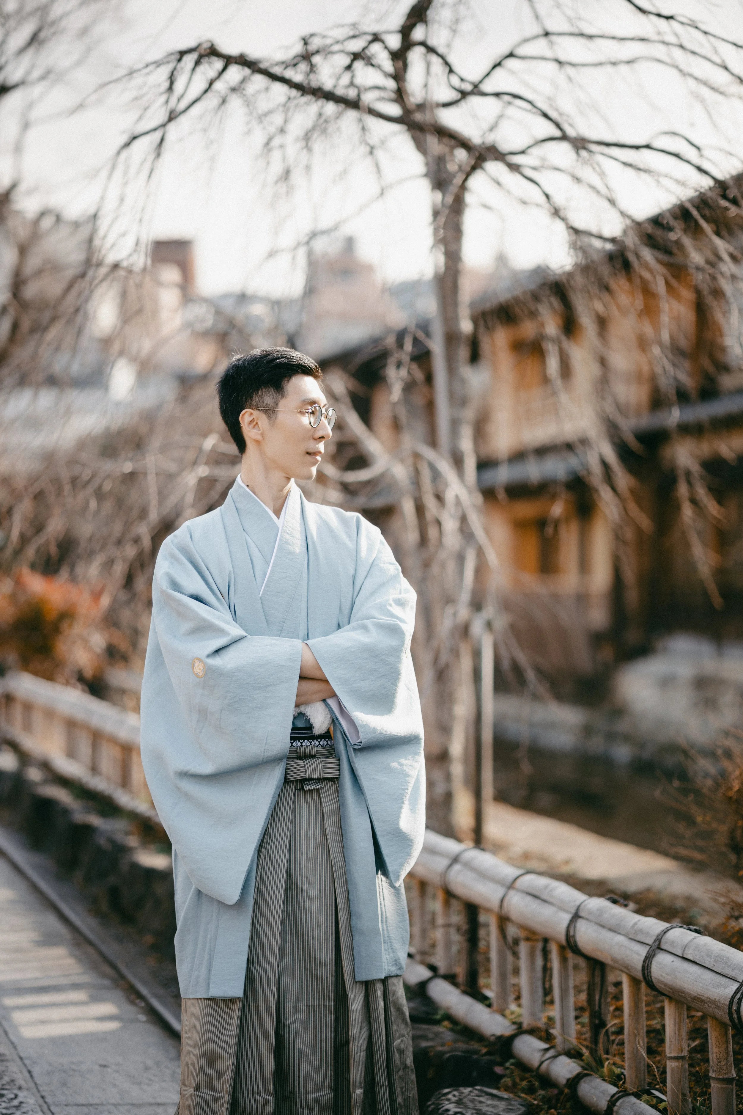 A young man in traditional Japanese clothing, including a light gray kimono and hakama, stands by a bamboo fence along a canal with bare trees and traditional wooden buildings in the background.