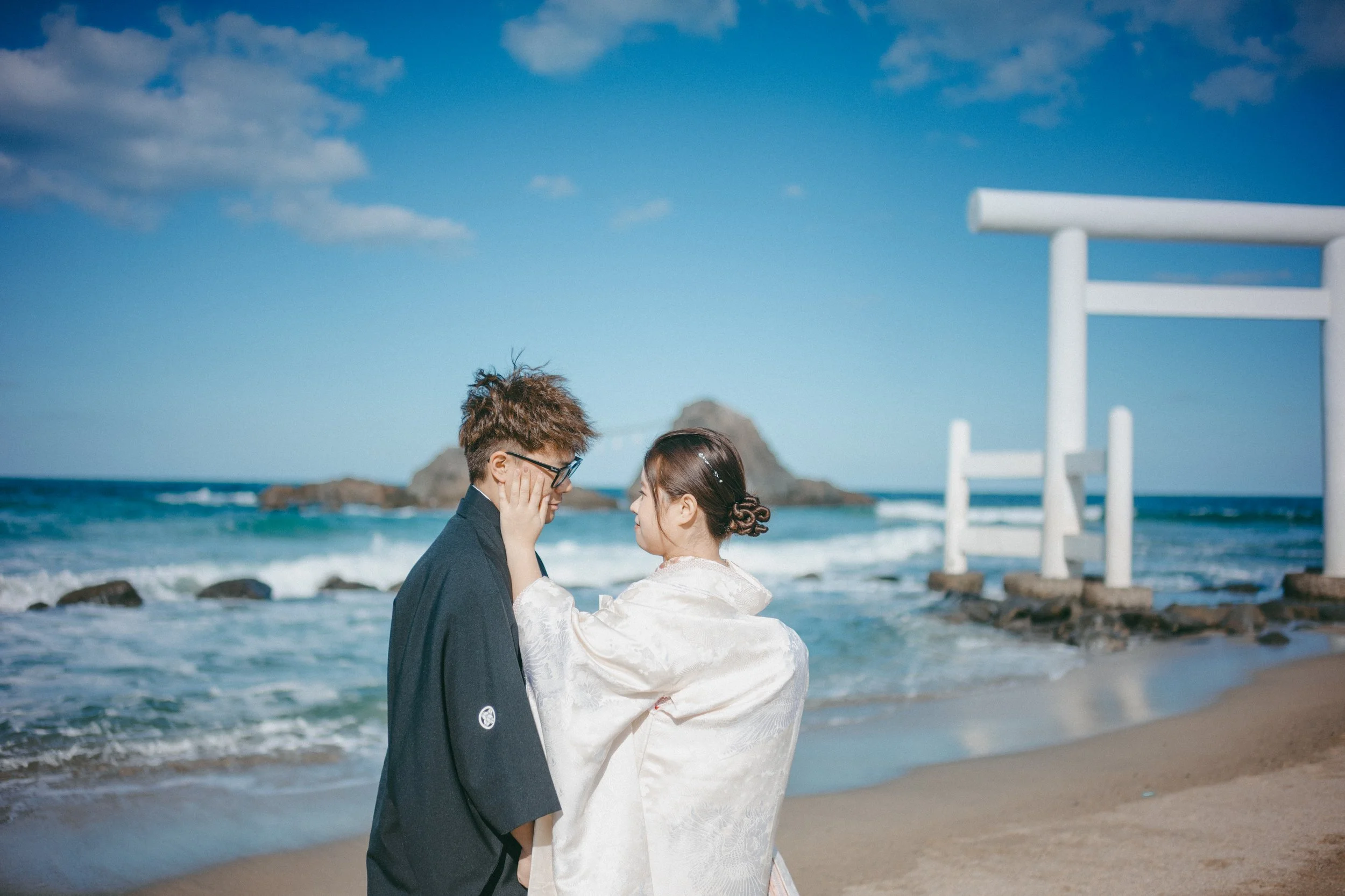 A couple dressed in traditional Japanese wedding attire stands on a beach near a white torii gate, with the ocean and rocky island in the background.