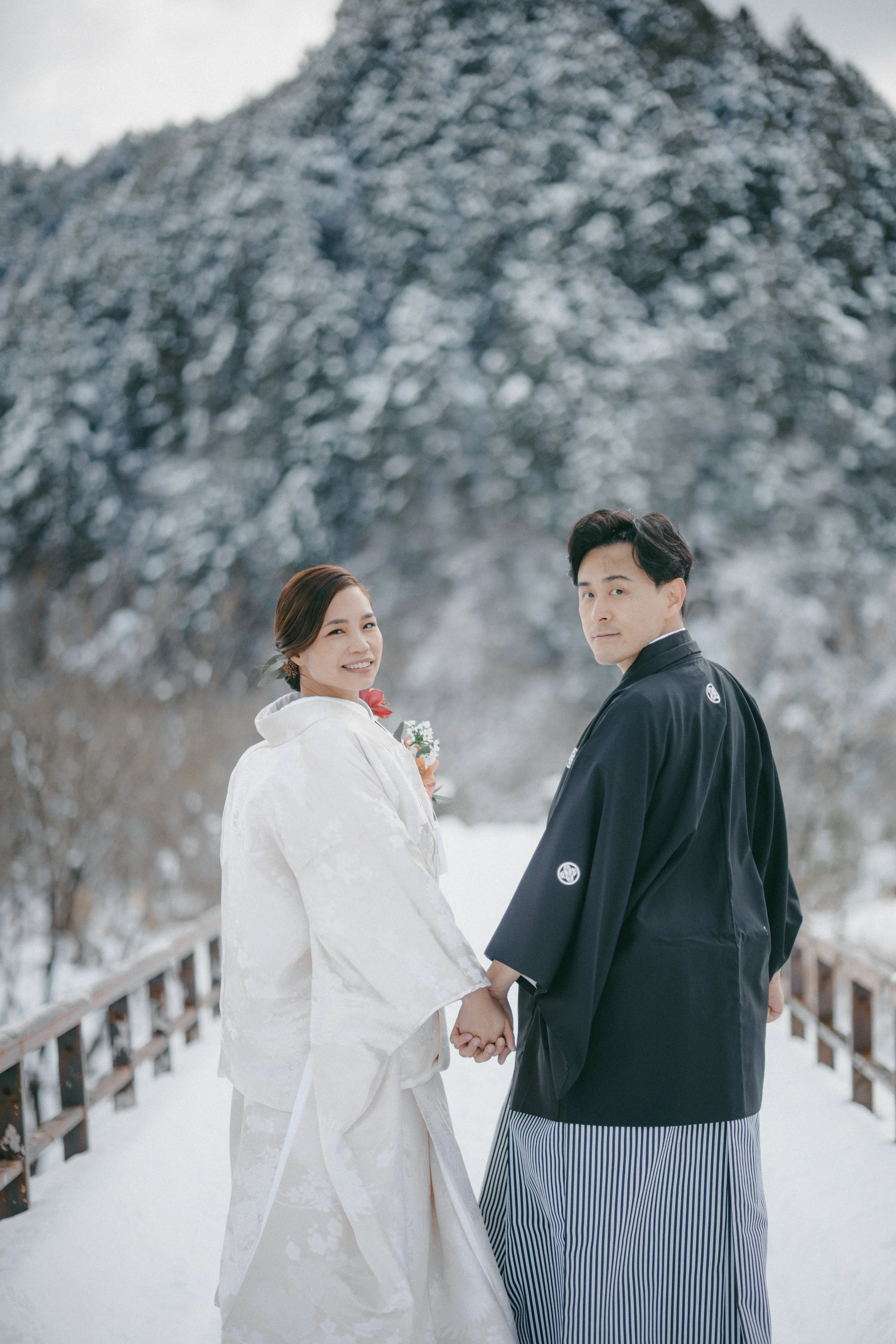 A Japanese couple in traditional wedding attire holding hands outdoors on a snowy day with snow-covered trees and mountain in the background.