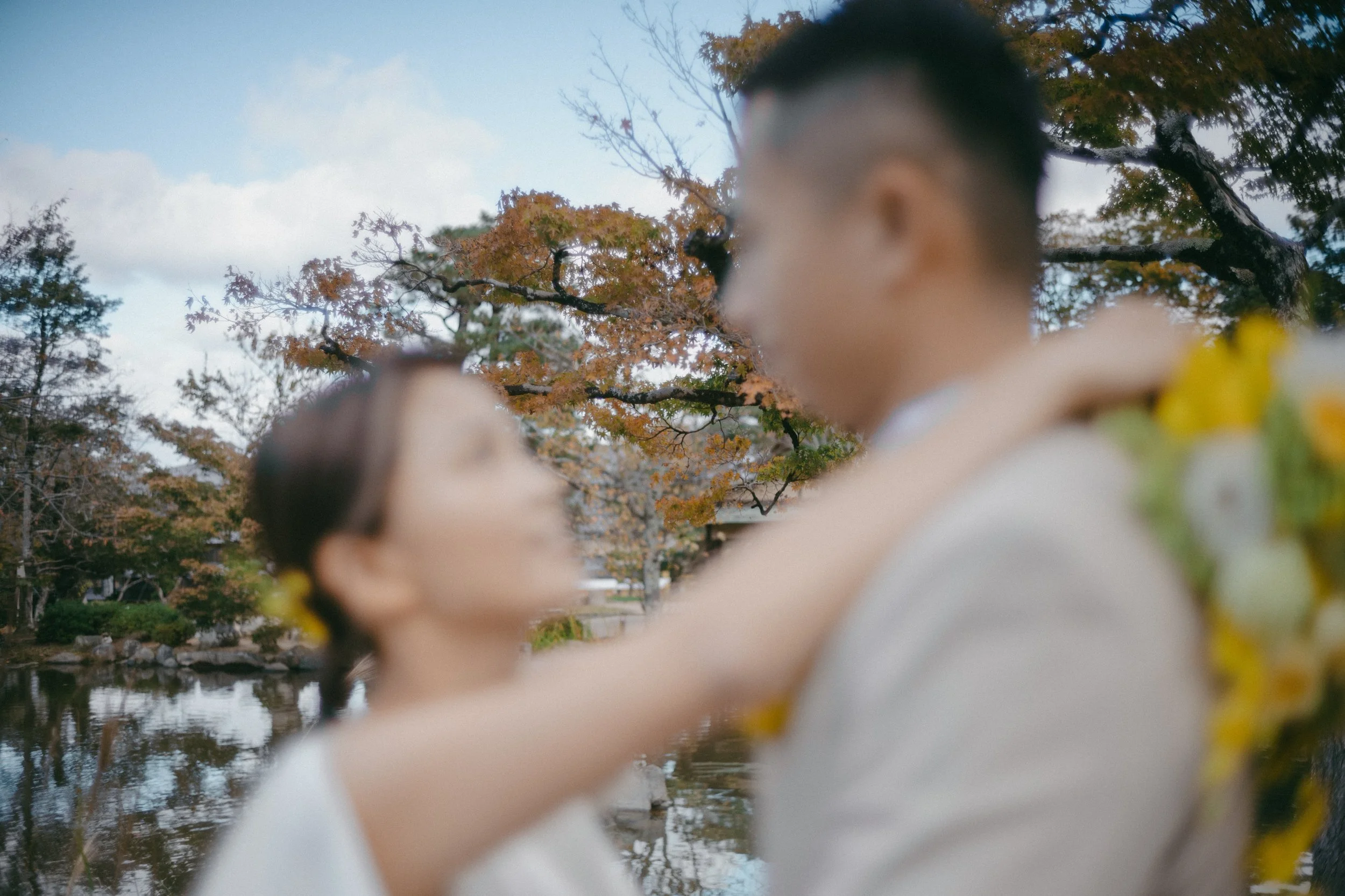 A couple standing close together outdoors by a lake with autumn trees in the background, blurred focus.