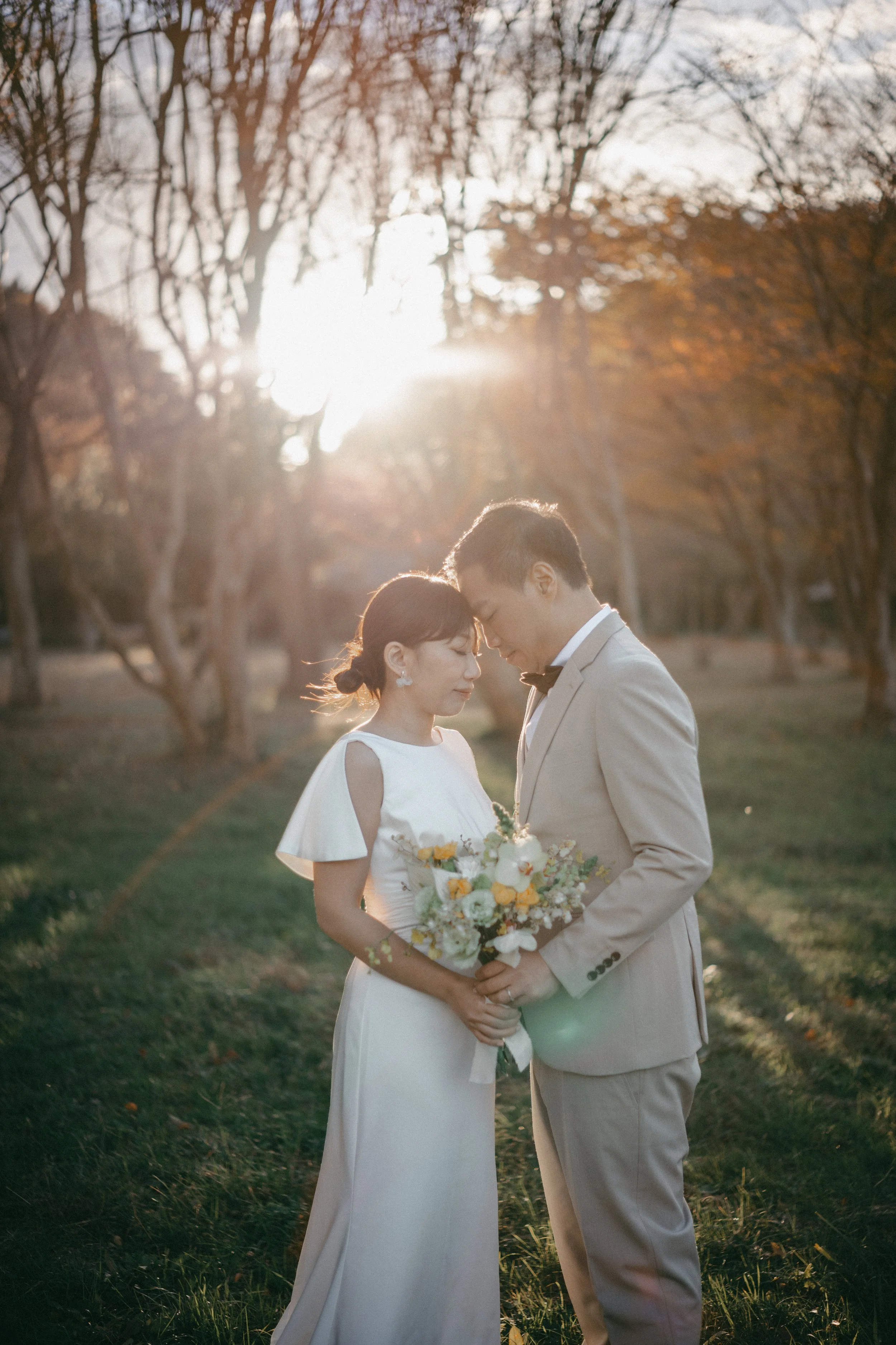 A bride and groom stand close with foreheads touching in a park during sunset, holding a bouquet of flowers.