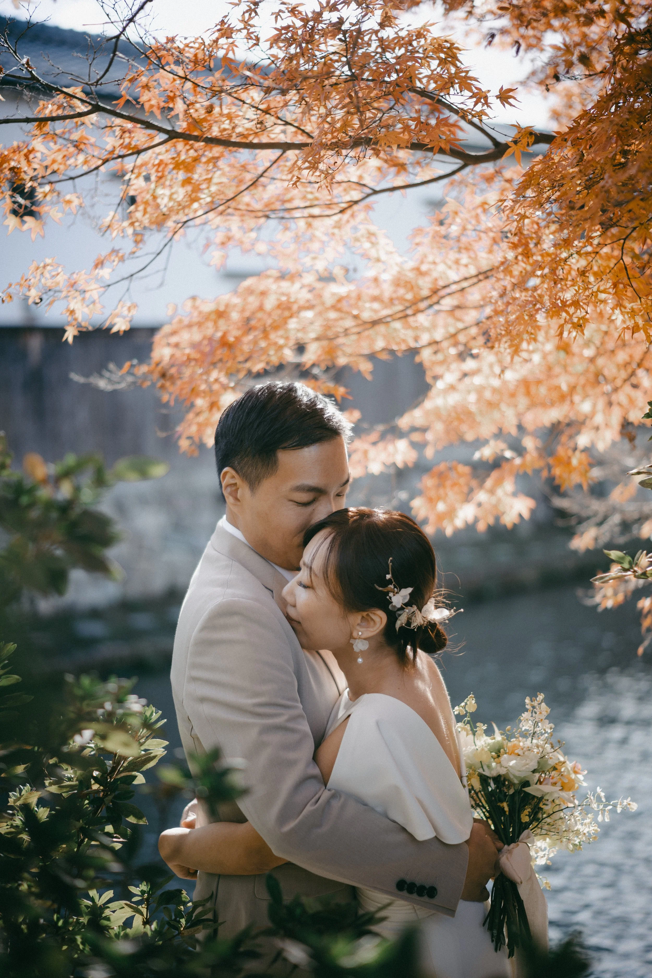 A bride and groom embrace outdoors under orange autumn leaves, with a river in the background. The bride holds a bouquet and has floral hair accessories, while the groom is in a light-colored suit.