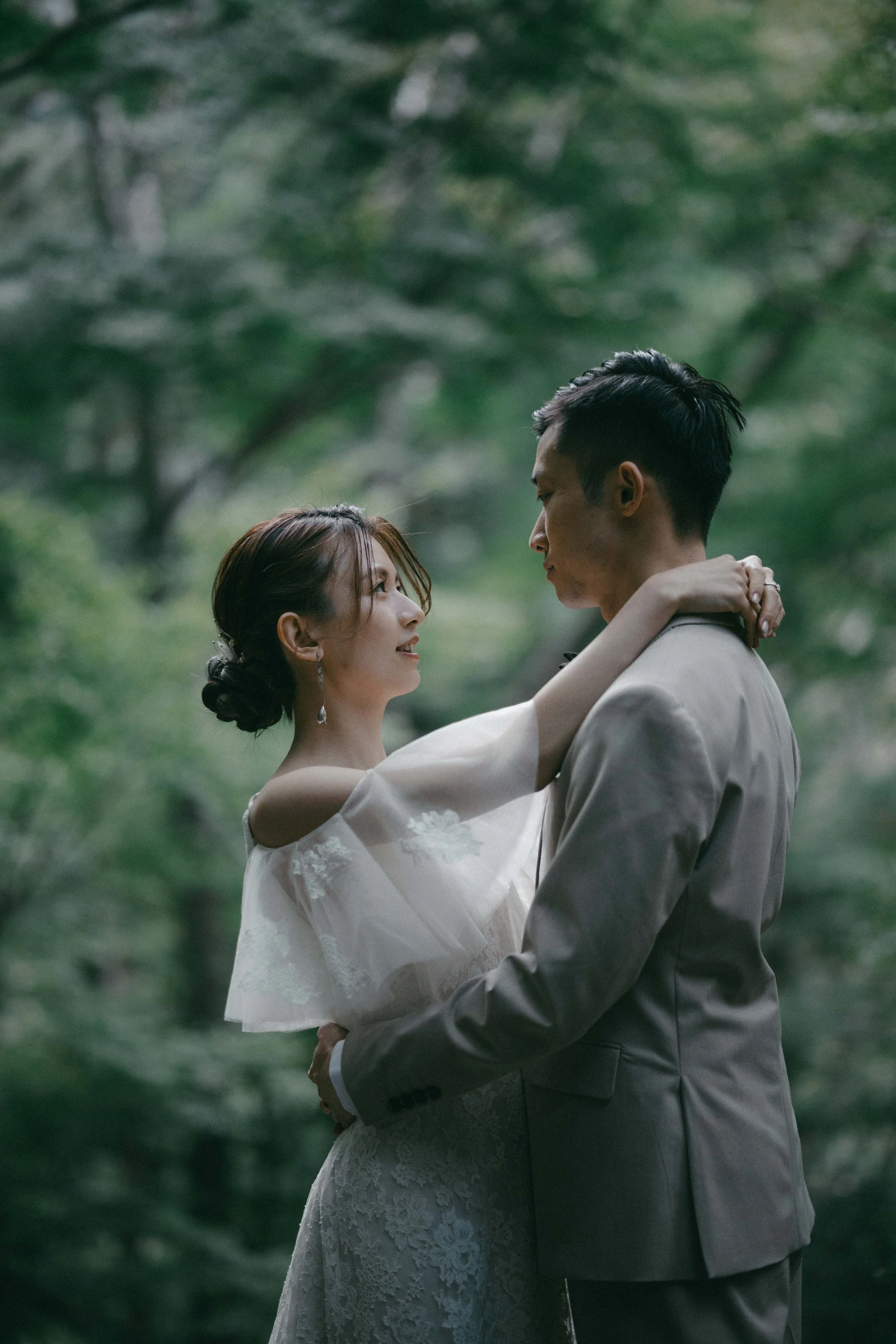 A bride and groom embrace in a forest, looking into each other's eyes.