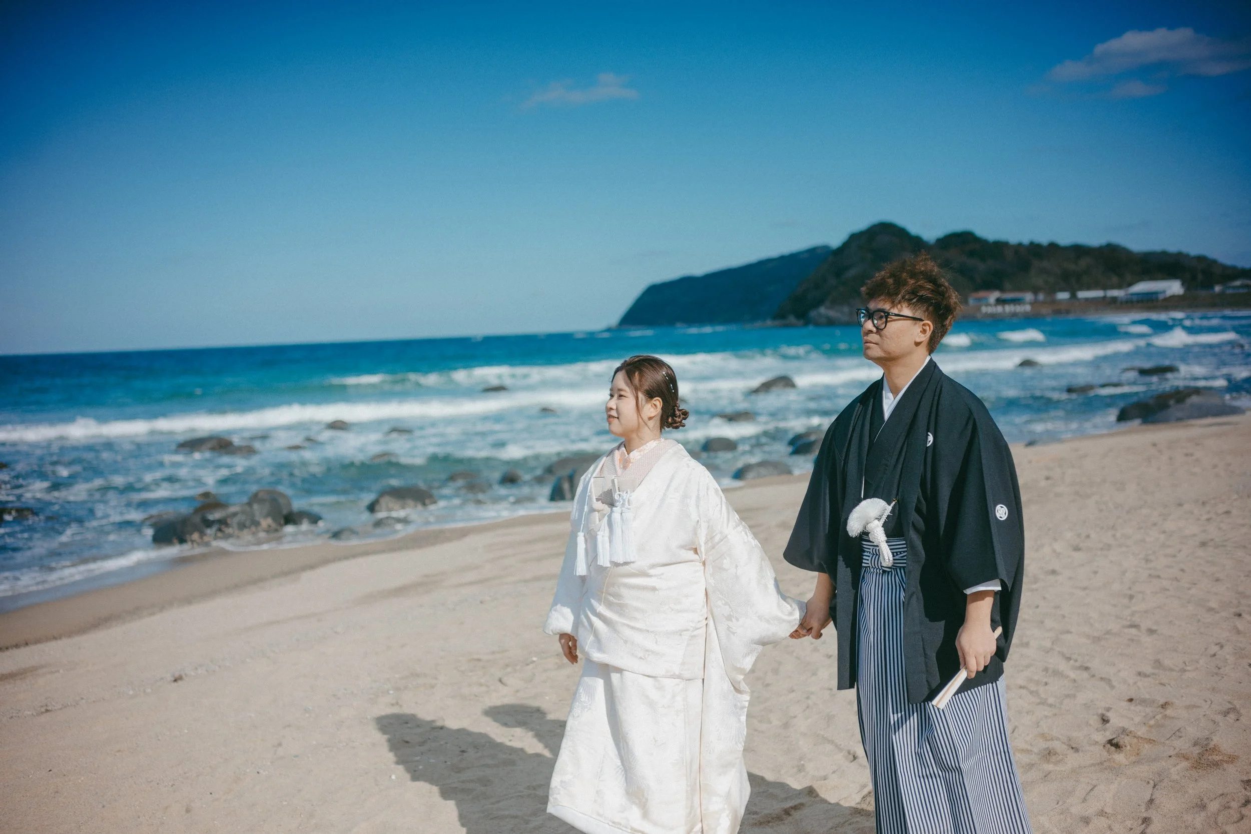 A young girl and a young man in traditional Japanese wedding attire walking hand in hand on a sandy beach with the ocean and mountains in the background.