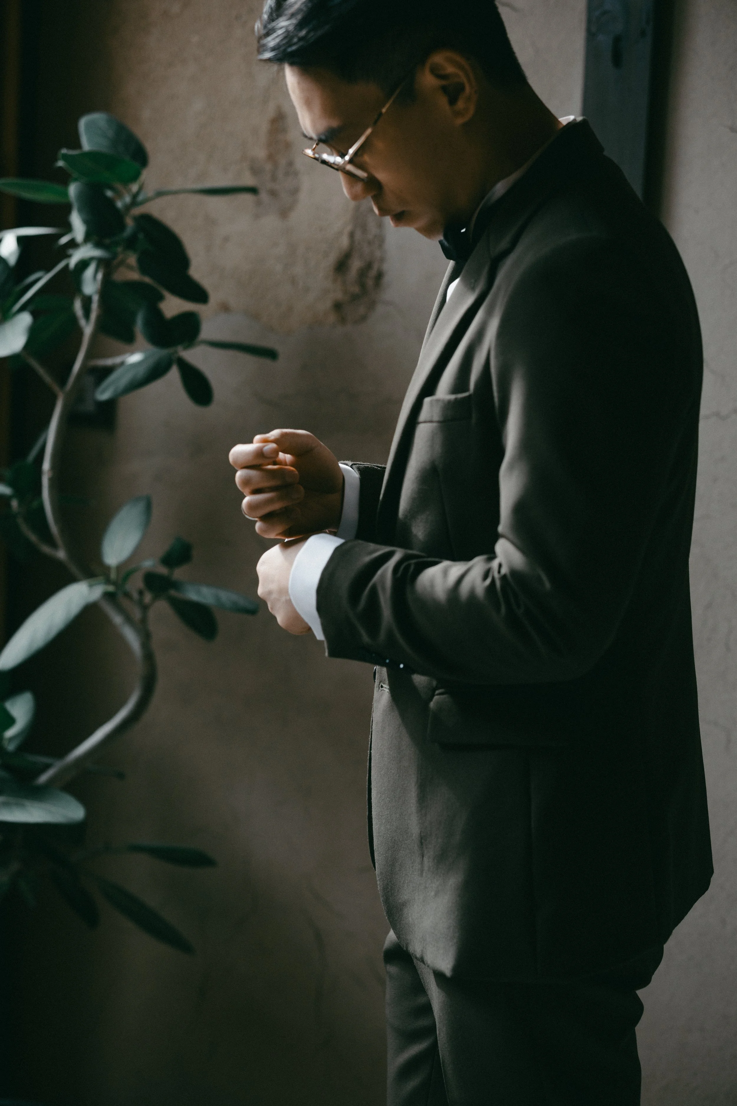 A man in a dark suit and glasses adjusting his cufflinks in a dimly lit room with a plant and textured wall in the background.