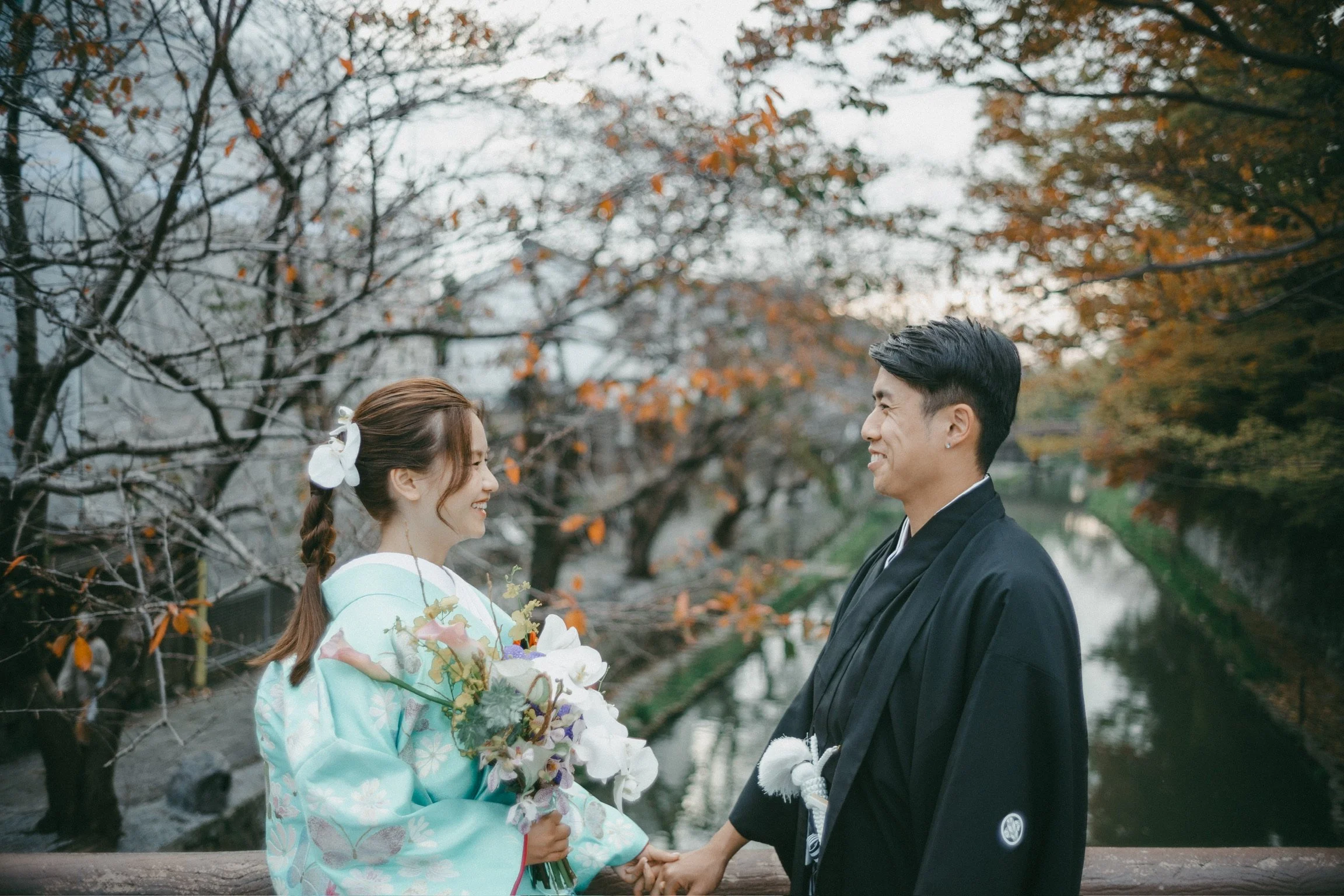 A young couple in traditional Japanese wedding attire standing on a bridge over a river, holding hands and smiling at each other, with autumn-colored trees in the background.