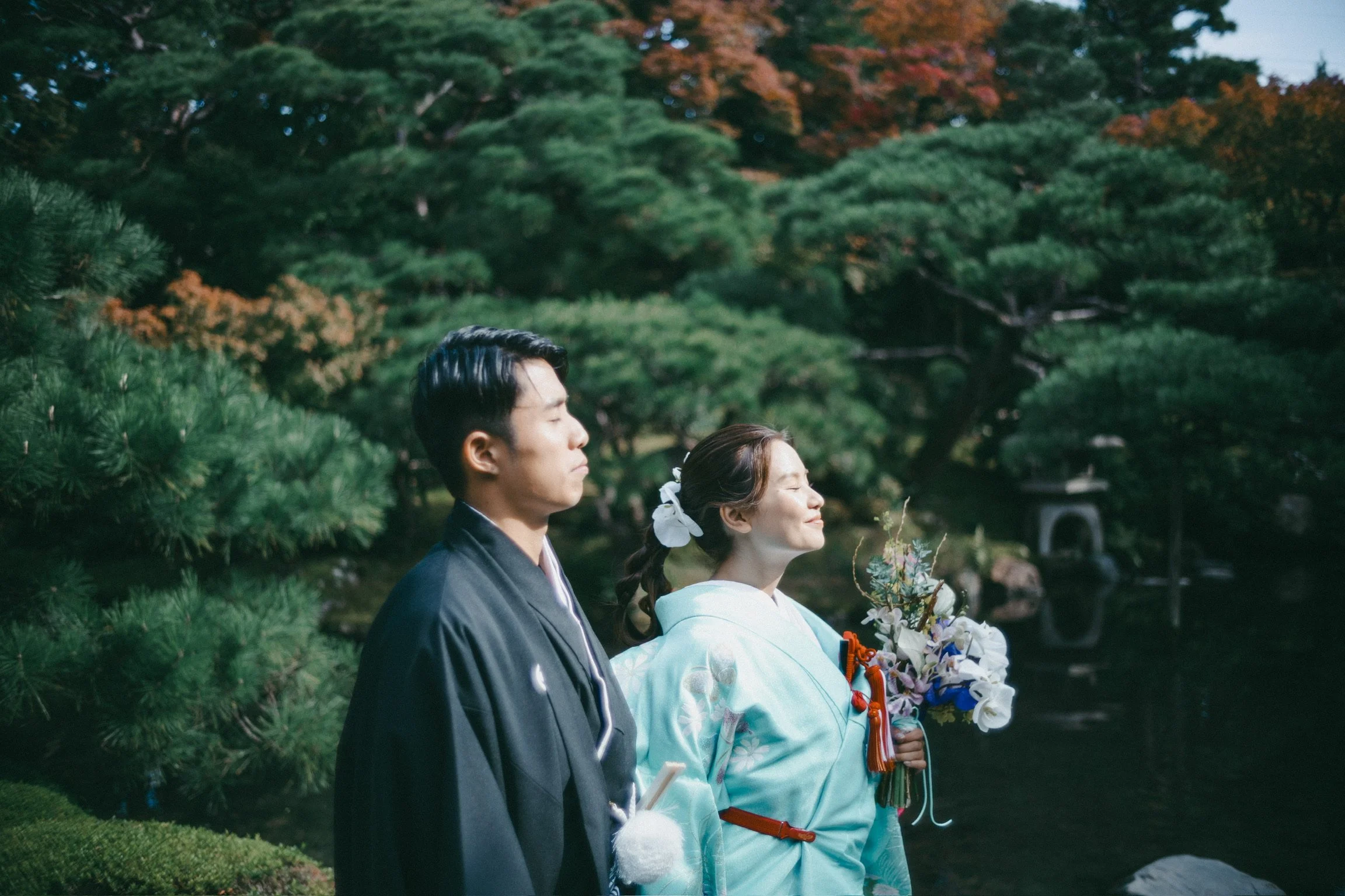 A couple in traditional Japanese wedding attire standing outdoors in a garden with greenery and a pond, the woman holding a bouquet of flowers.