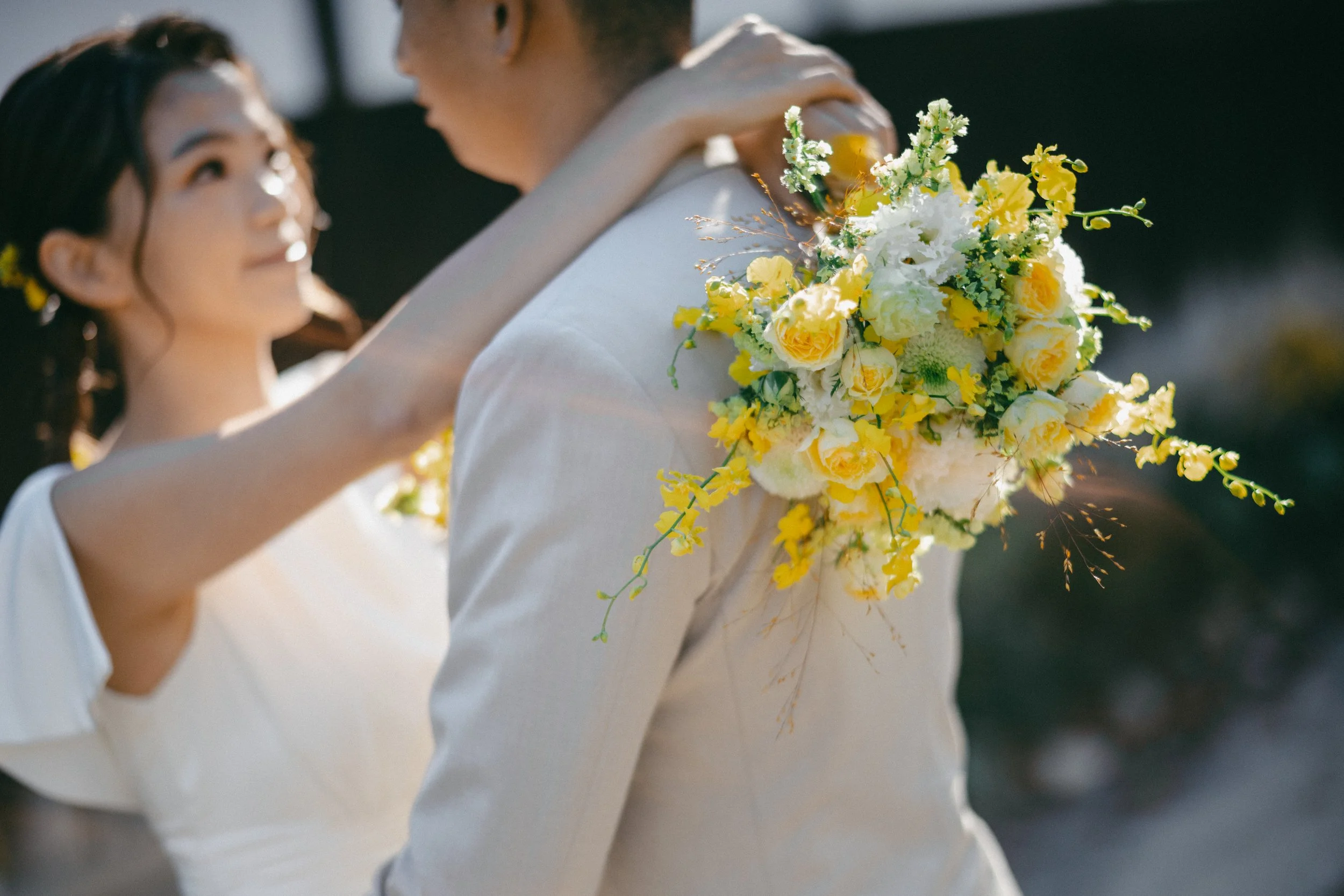 A bride and groom sharing a moment, the bride with her hand on the groom's shoulder, holding a bouquet of white and yellow flowers, with the groom's back turned.