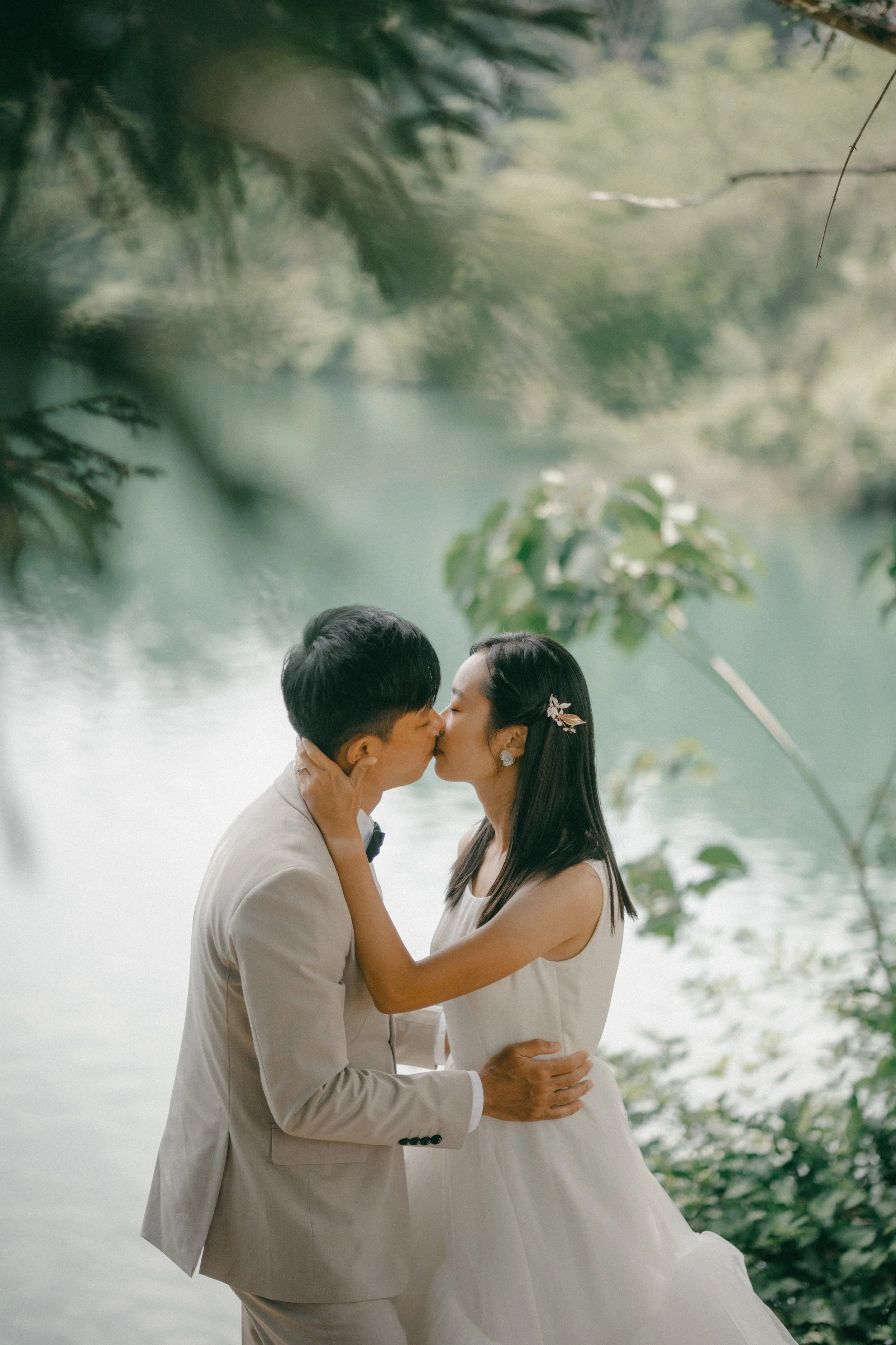 A couple in wedding attire sharing a kiss by a lakeside surrounded by greenery.