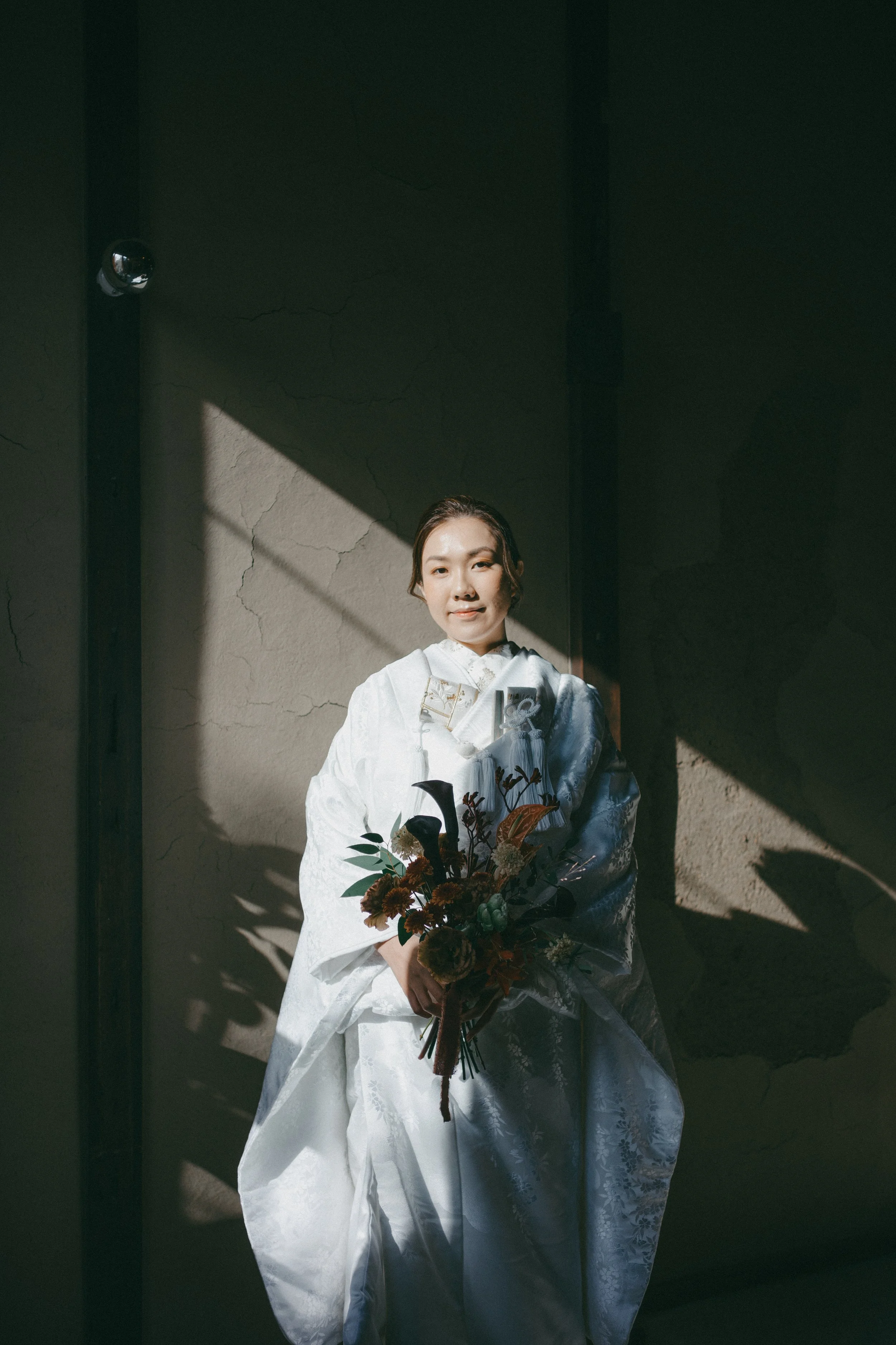 A woman in a white traditional wedding dress holding a bouquet of flowers, standing against a wall with sunlight casting shadows.