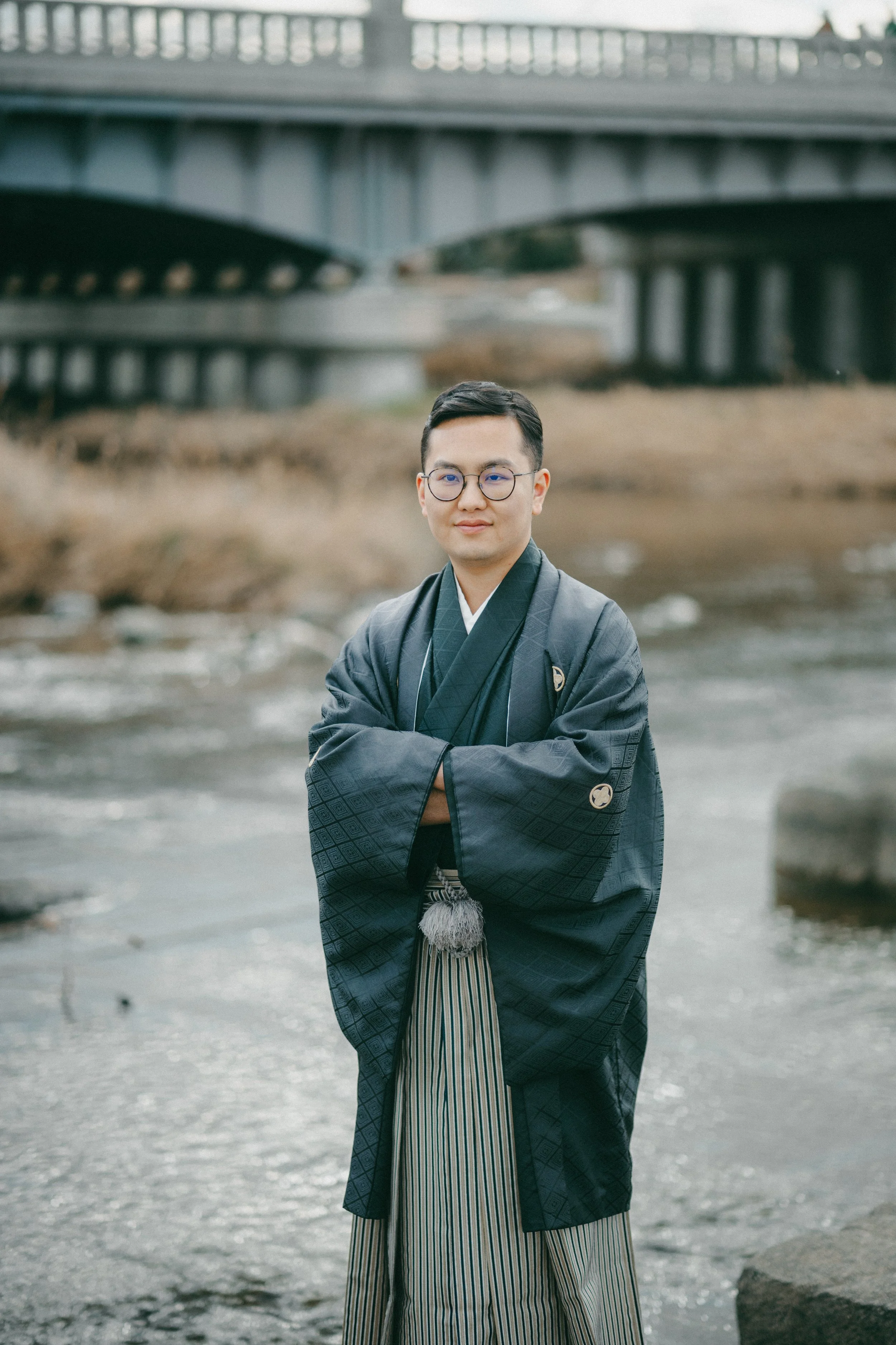 A man in traditional Japanese attire standing near a river with bridges in the background.