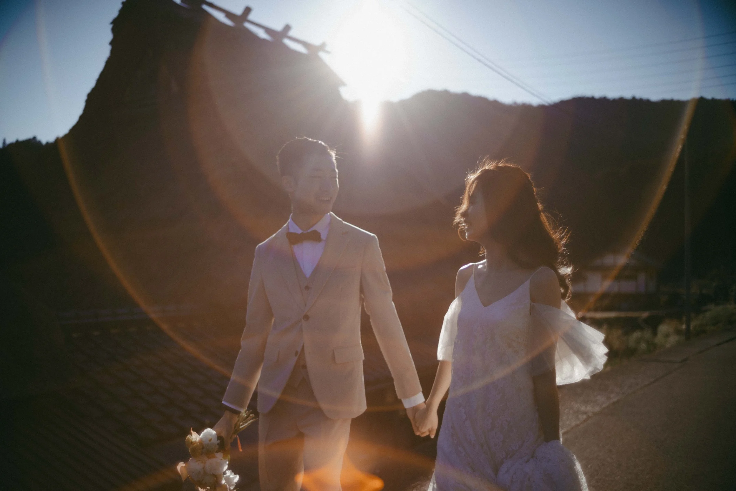 A couple holding hands outdoors during sunset, with a scenic background of hills and power lines. The man is wearing a beige suit with a black bow tie, holding a bouquet, and the woman is wearing a white lace dress with ruffled sleeves.