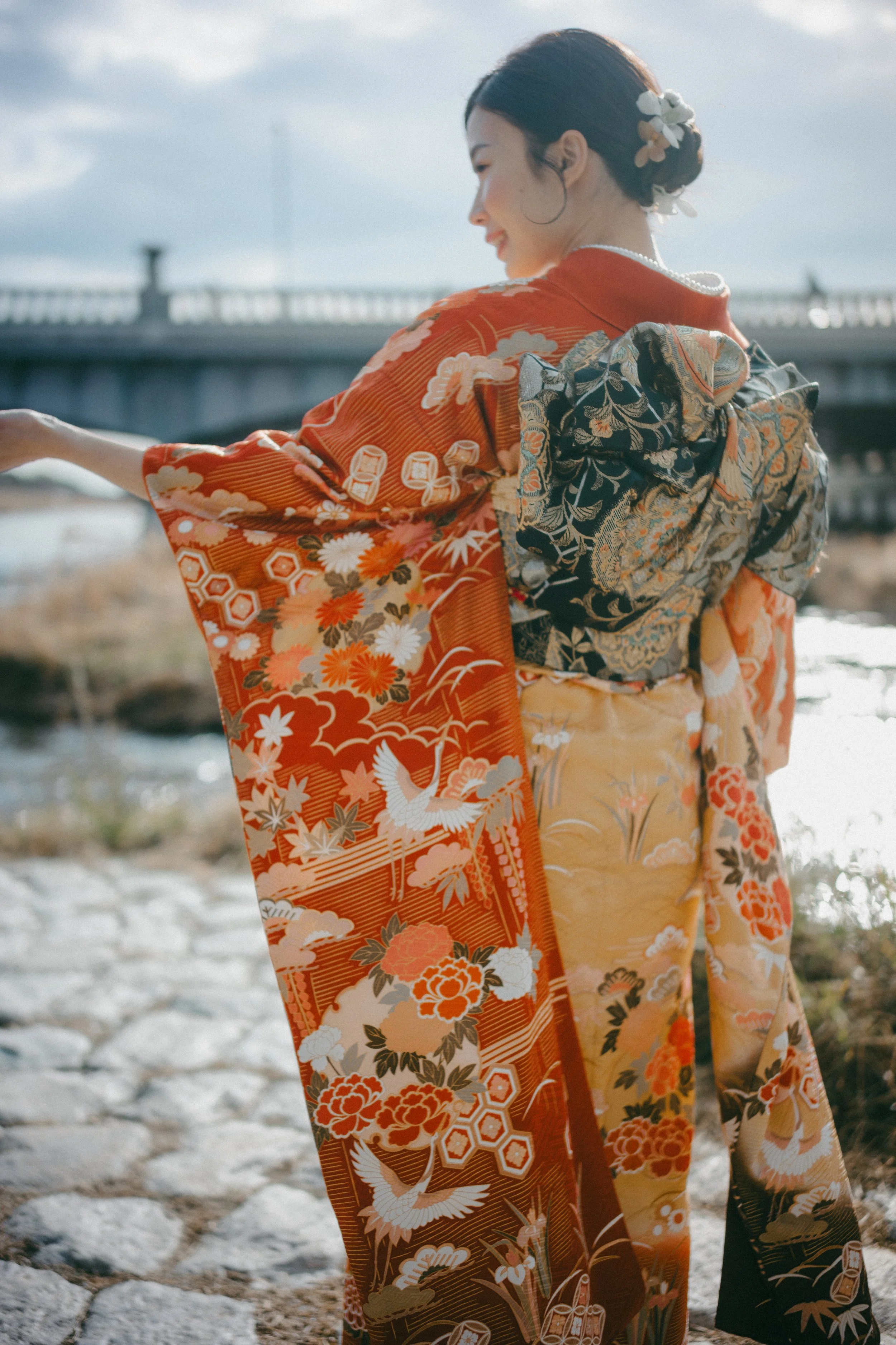 A woman wearing a colorful, traditional Japanese kimono with floral and nature patterns, standing outdoors near a river and a bridge, smiling.