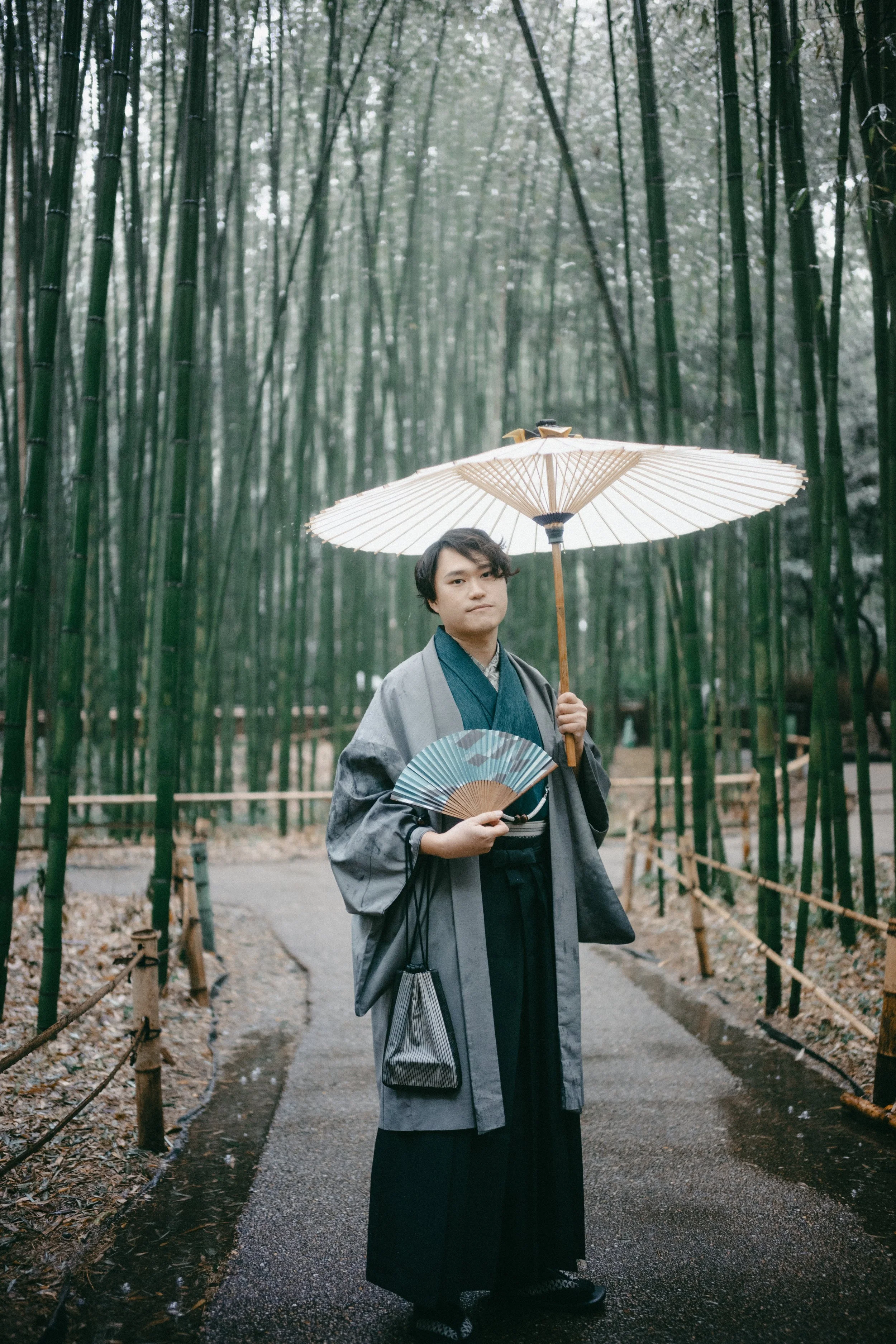 A young person in traditional Japanese attire holding a paper fan and an umbrella, standing on a path in bamboo forest.