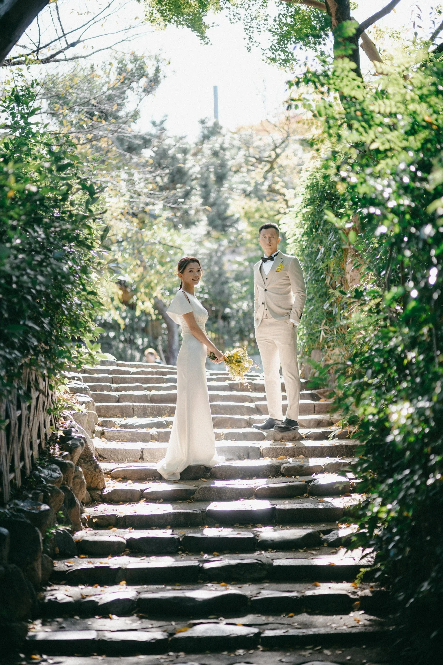 A bride and groom standing on stone steps outdoors, surrounded by green trees, with sunlight filtering through the leaves, in a wedding setting.