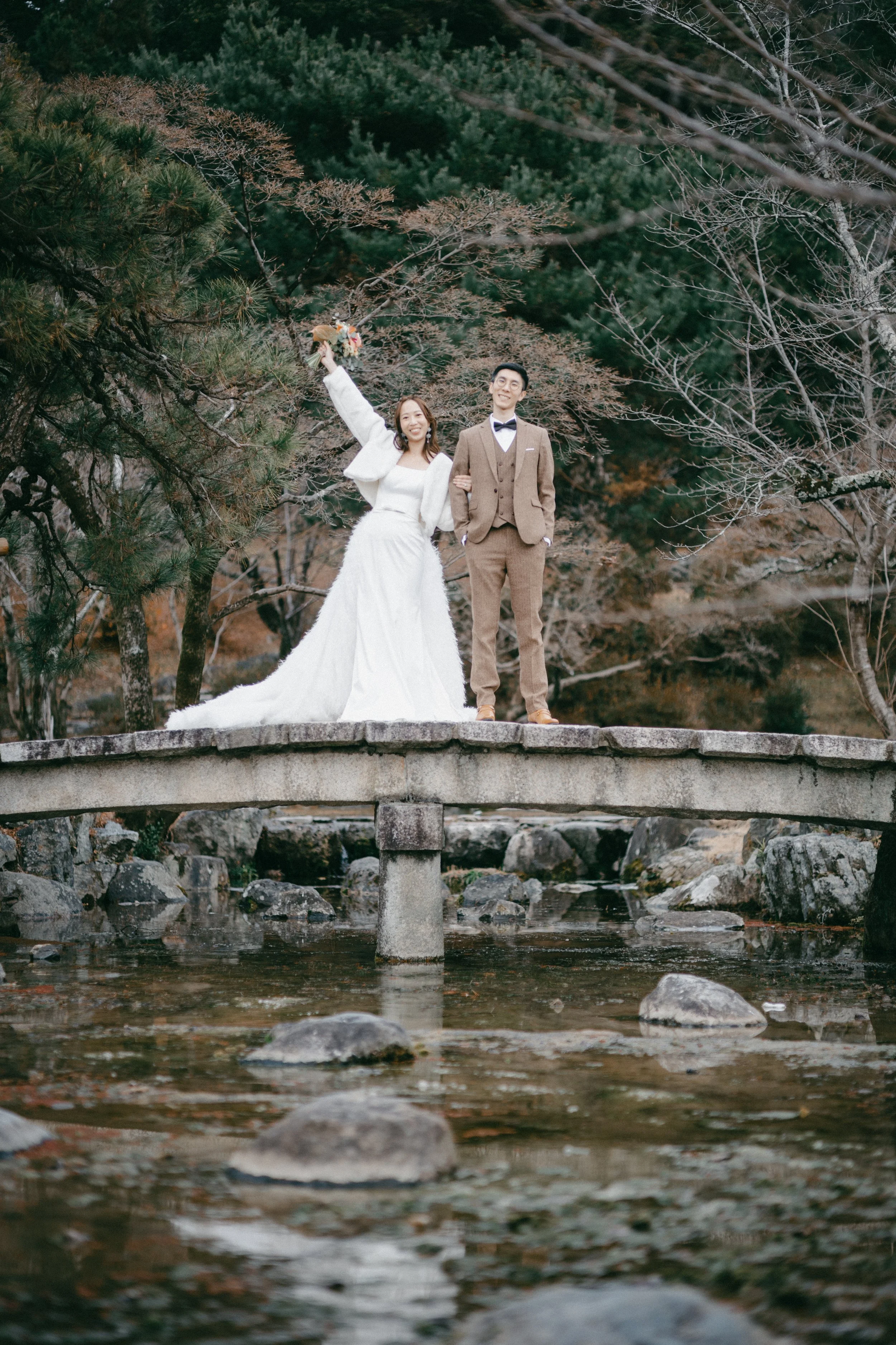 A newlywed couple standing on a stone bridge over a stream surrounded by trees with autumn foliage. The bride is smiling and holding a bouquet of flowers with her arm raised; she wears a white wedding dress and matching jacket. The groom stands besid