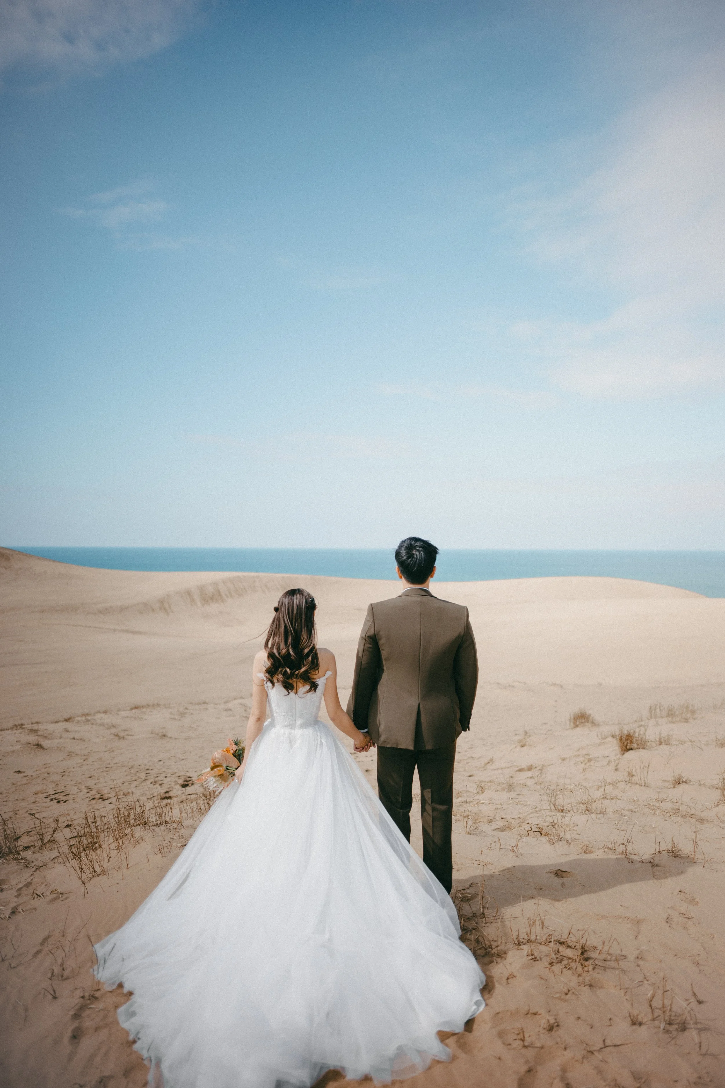 A bride in a white wedding dress and a groom in a dark suit walking hand in hand through sandy dunes towards the ocean under a partly cloudy sky.